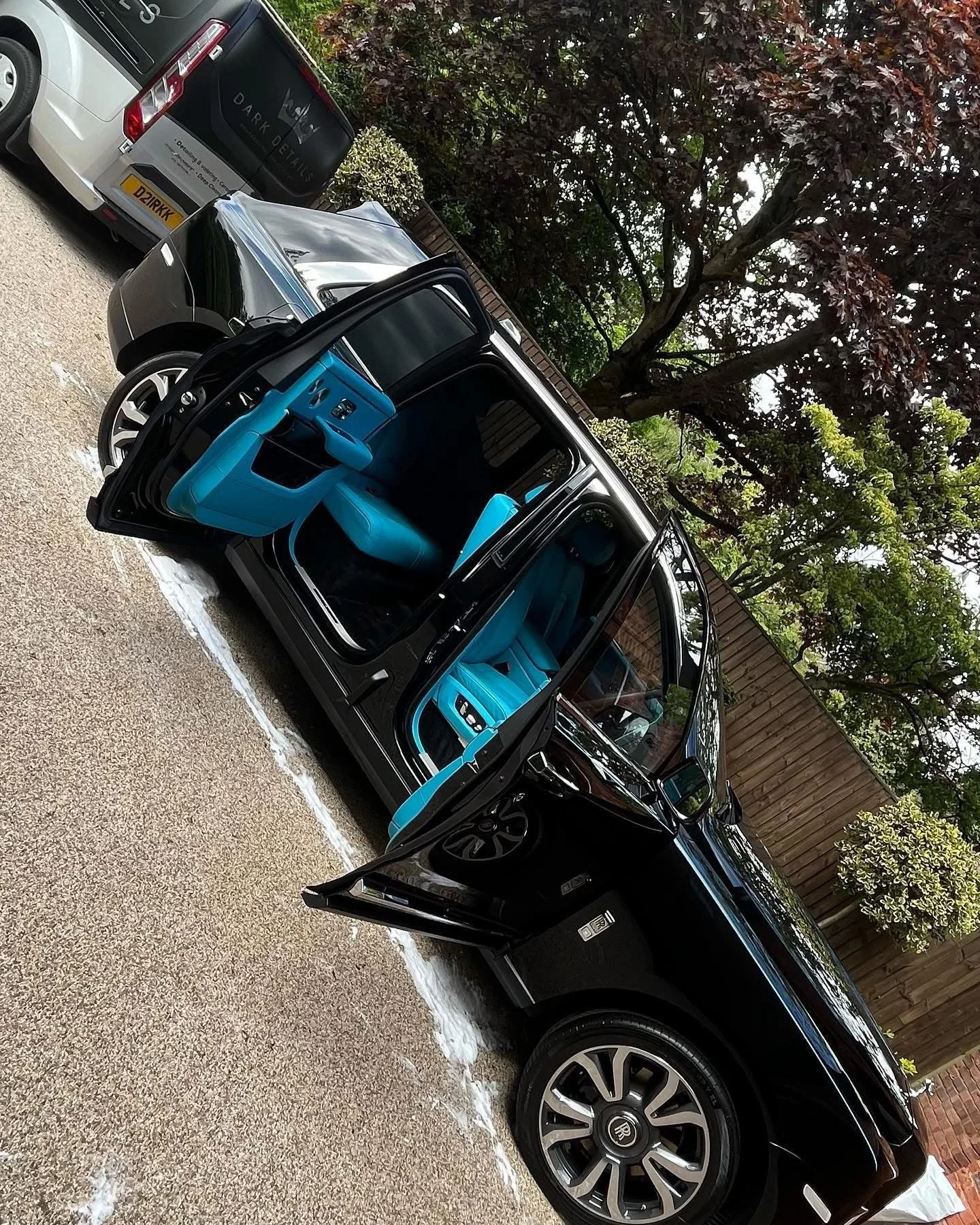 A black compact car with open driver's side door, revealing bright blue interior seats and dashboard, parked on a pebble driveway with other cars and trees in the background.