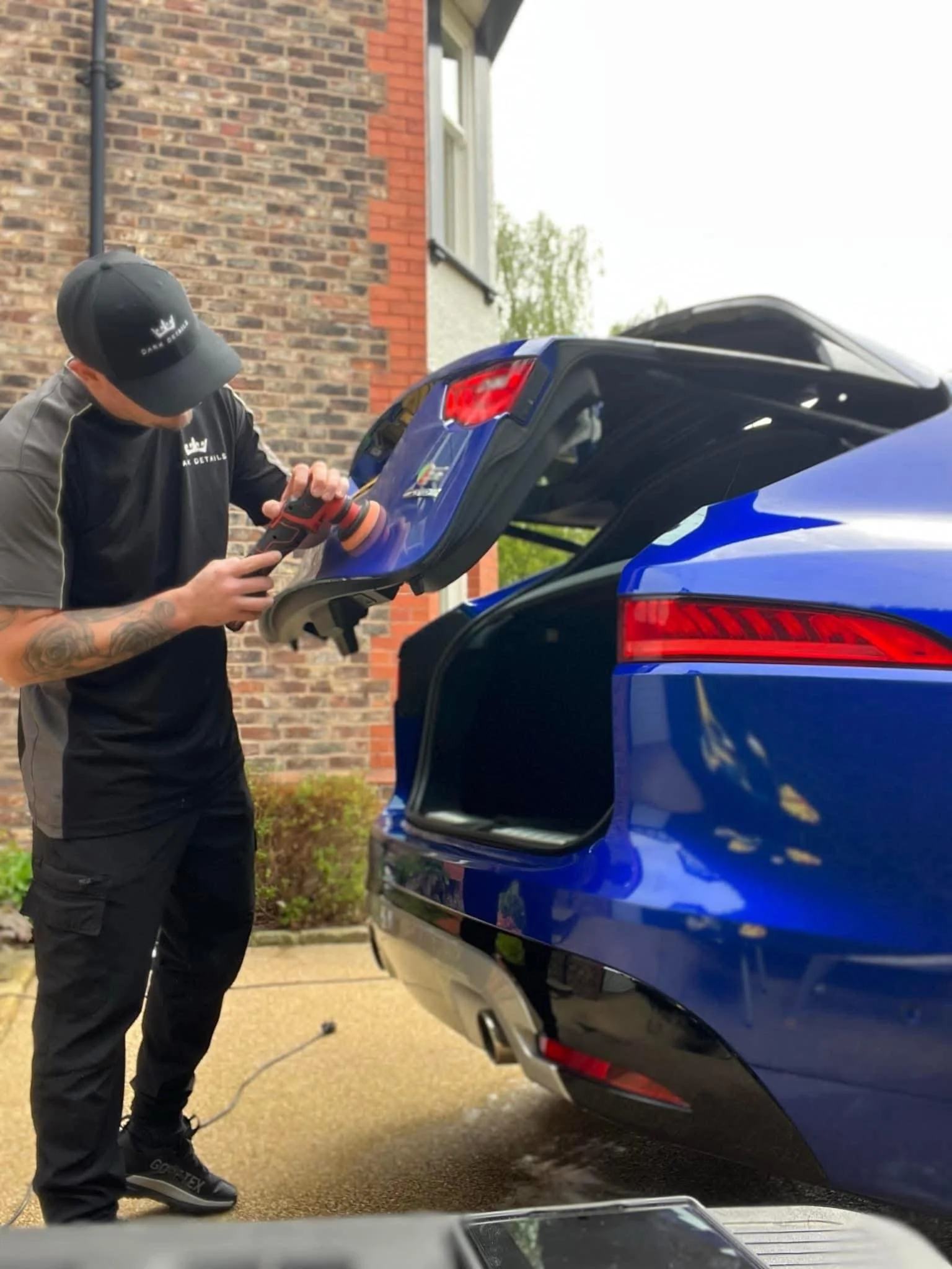 A person wearing a black cap and black shirt is using a machine to polish or clean the trunk of a blue car. The car's trunk is open, and the person is focused on the task. The background shows a brick wall and some greenery.