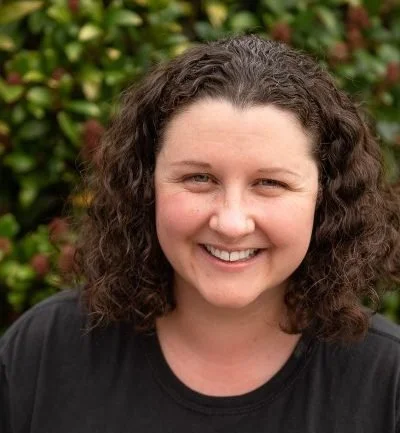 Nicki Cawood, Journal therapist and copywriter, a woman with curly brown hair smiling outdoors with green and red foliage in the background.