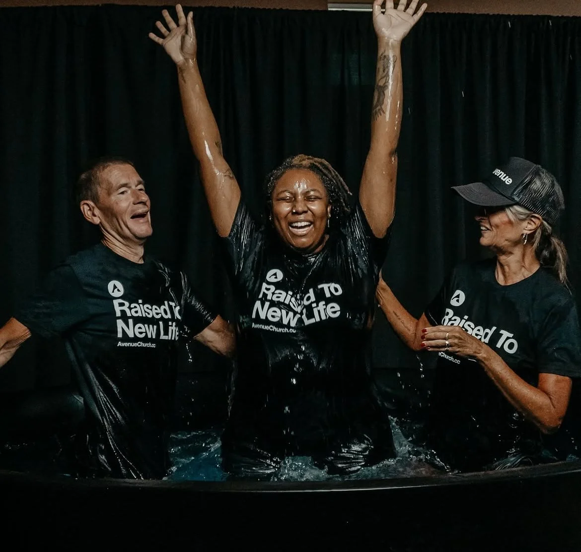 Three women in black shirts with the text "Raised To A New Life" are celebrating in a baptismal pool, with the central woman raising her arms and smiling, flanked by two women smiling and touching her shoulders, against a black curtain background.