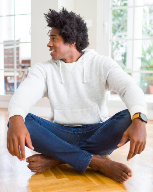 Young man sitting cross-legged on the floor in a bright room, wearing a white hoodie and blue jeans, smiling and looking to the side.