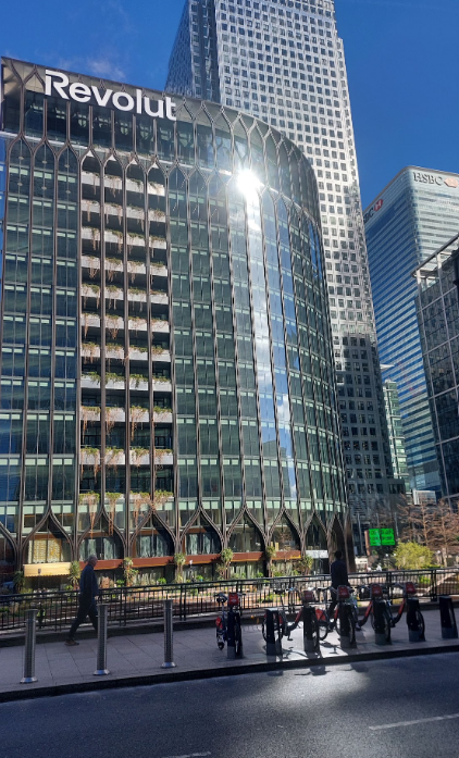 City street scene with tall modern office buildings, including the Revolut building, under a clear blue sky. Pedestrians and bikes are visible on the sidewalk.