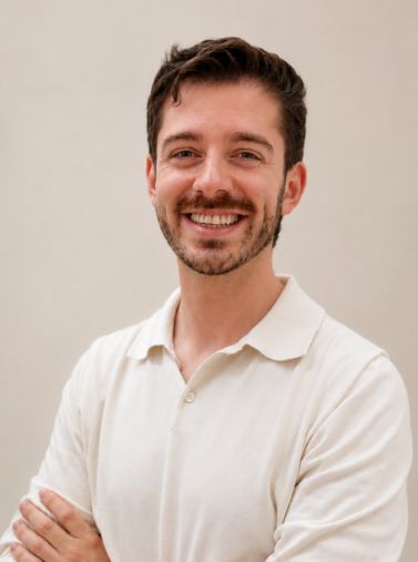 Portrait of George Brassil with brown hair and beard, smiling, wearing a white polo shirt, standing against a neutral background.