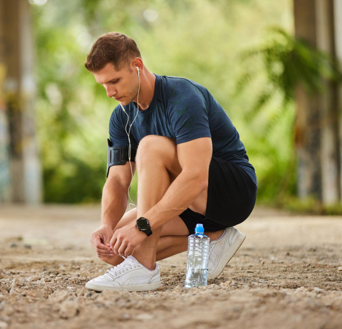 A man crouching outdoors tying his shoelace, wearing a navy athletic shirt, black shorts, white sneakers, and earphones, with a water bottle nearby.