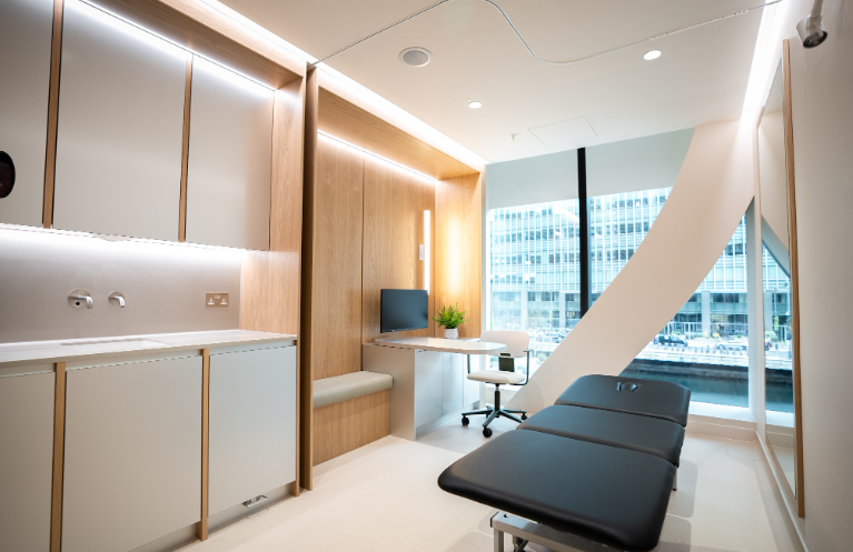 A modern medical examination room with a black examination table, a small white desk with a computer monitor and a potted plant, and large windows with city views.
