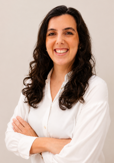 Smiling Sara Pedro Dias with long, curly dark hair wearing a white shirt, crossing her arms, against a neutral background.