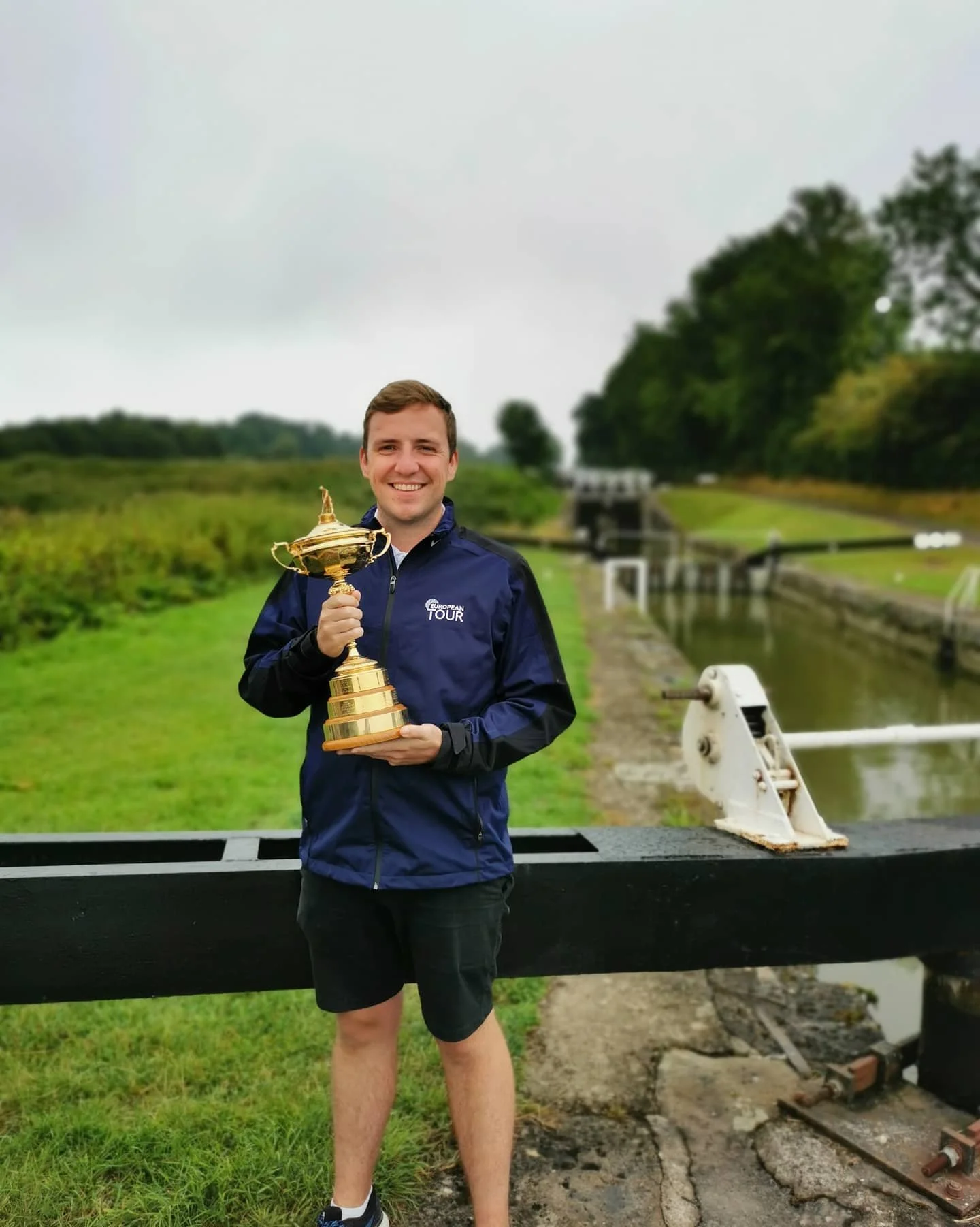 A man smiling and holding a gold trophy in an outdoor setting with a canal and greenery in the background.