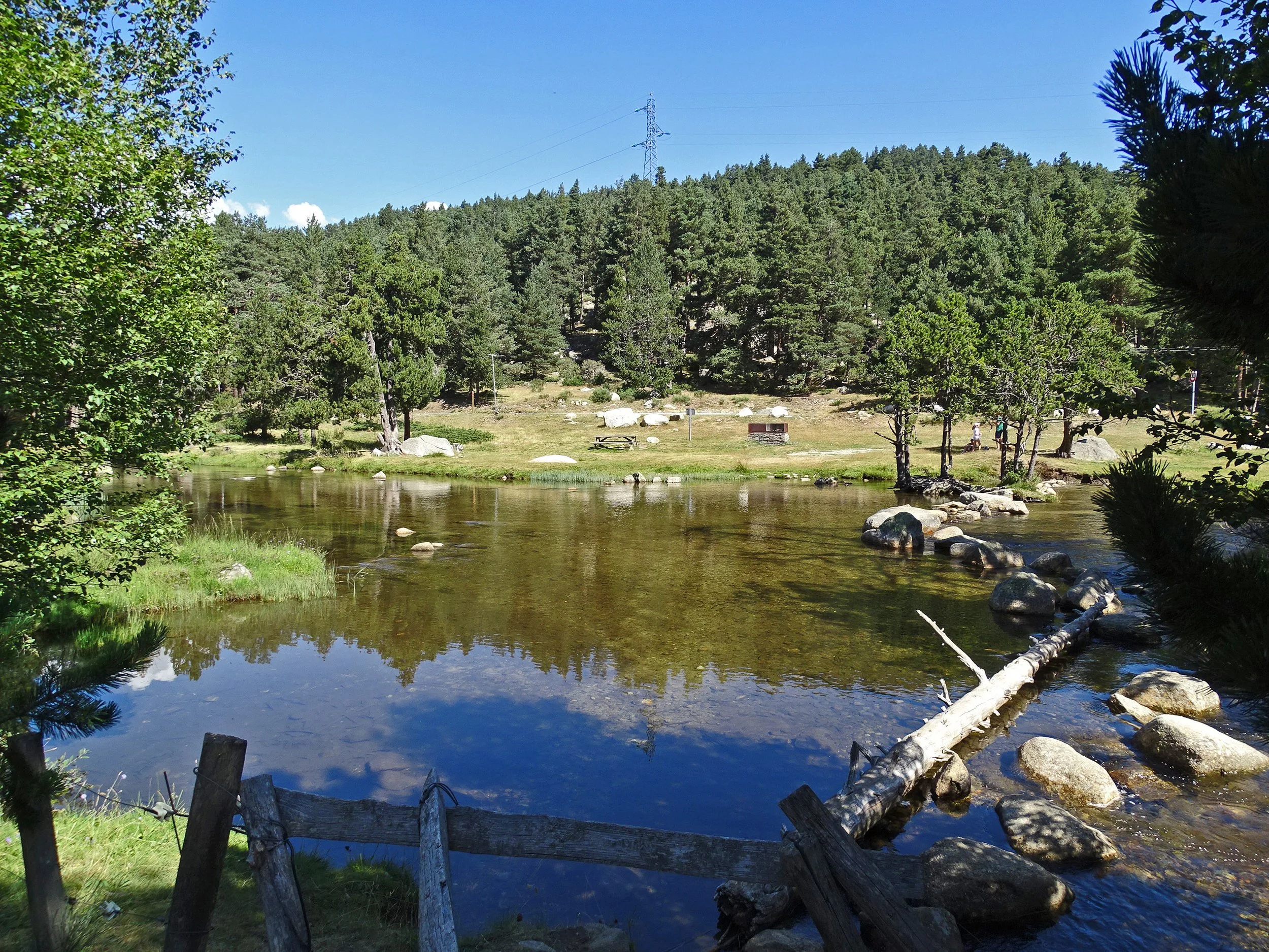 Une rivière calme entourée d'arbres verts et d'une forêt dense. Le ciel est bleu avec quelques nuages. Il y a une petite zone de pique-nique avec une table en bois et un barbecue en pierre. Quelques personnes marchent au fond de la scène.