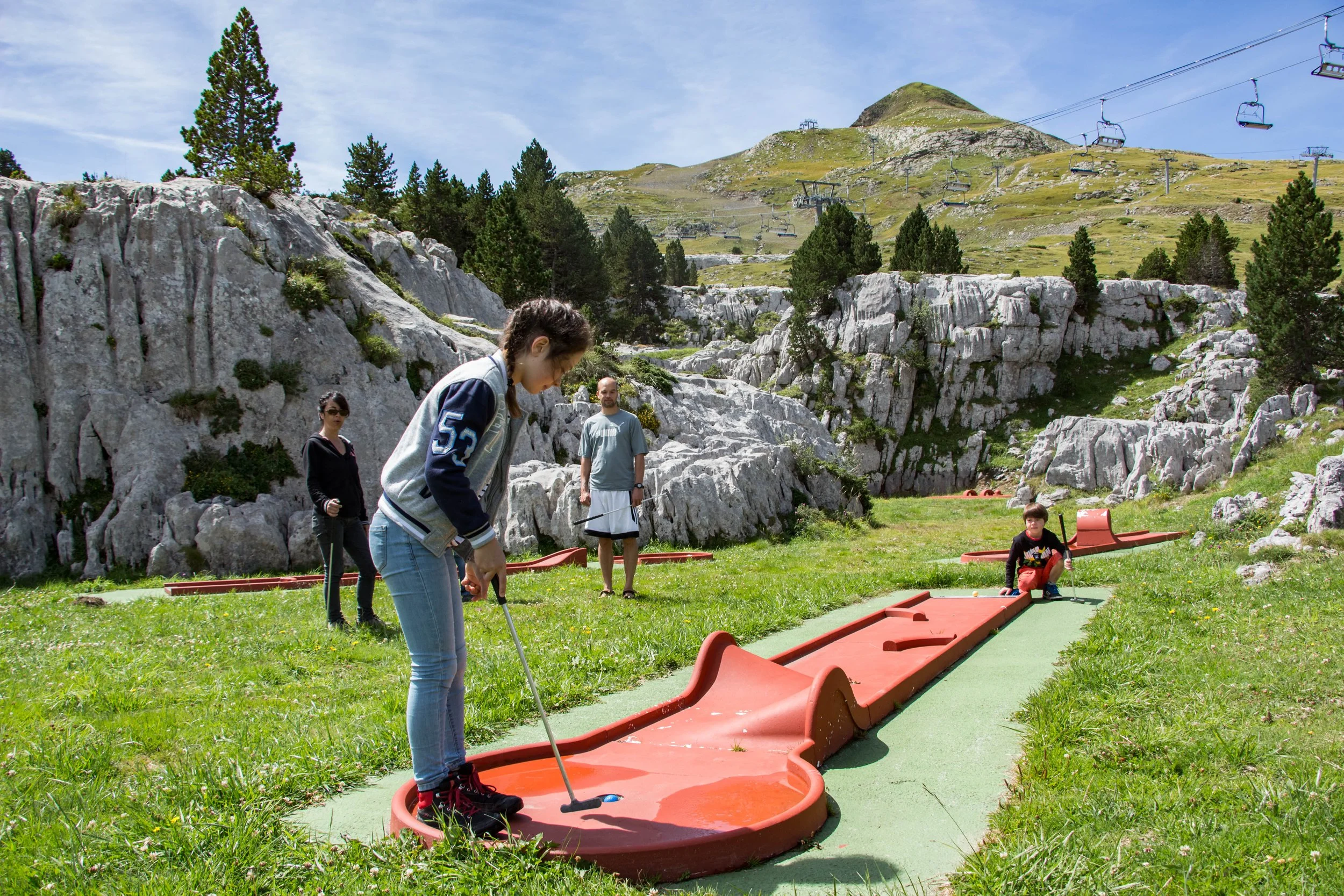 Groupe de personnes jouant au mini-golf en pleine nature avec une montagne et des sapins en arrière-plan.