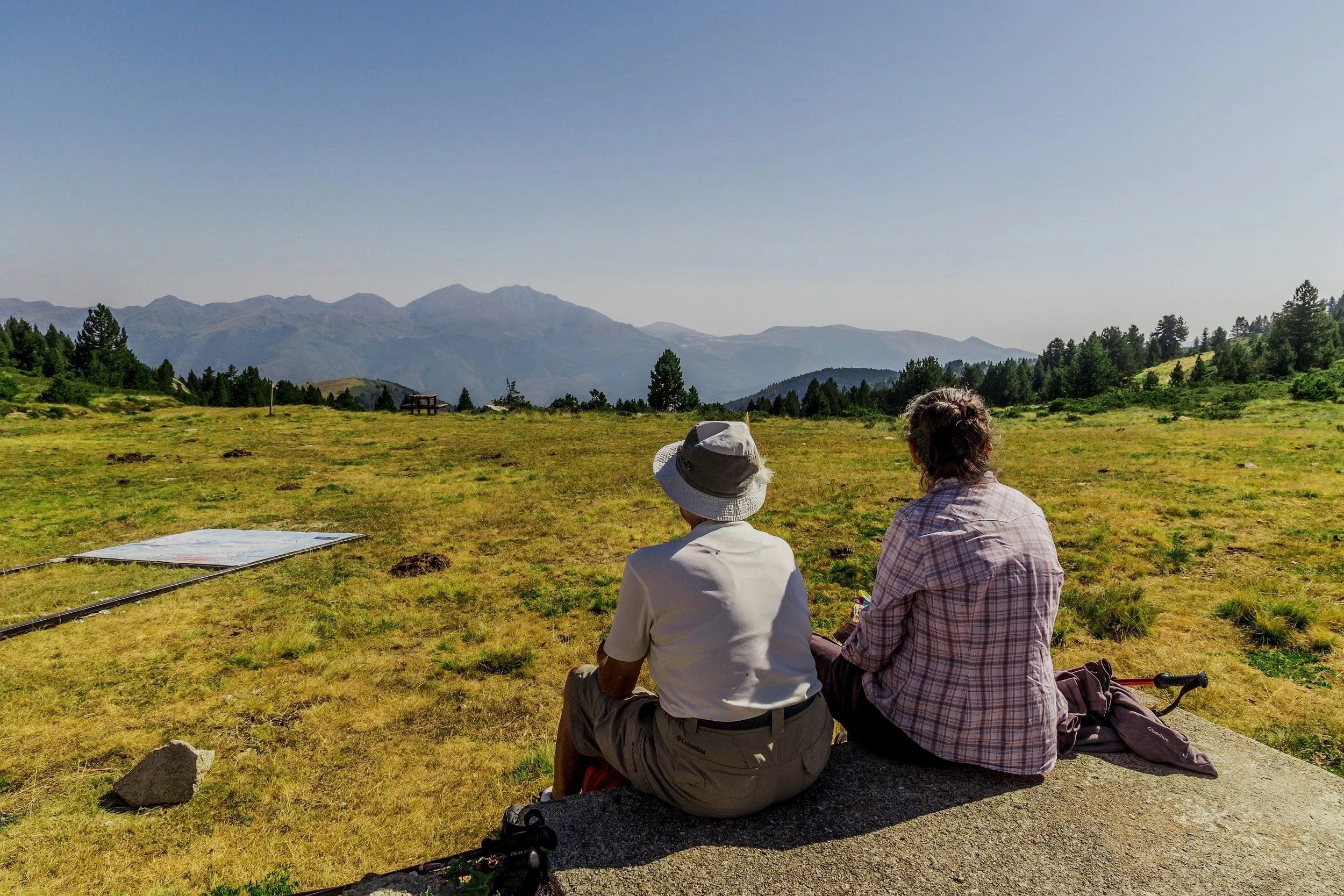 Deux personnes assises dans un champ verdoyant, regardant des montagnes au loin, avec un ciel clair et ensoleillé.