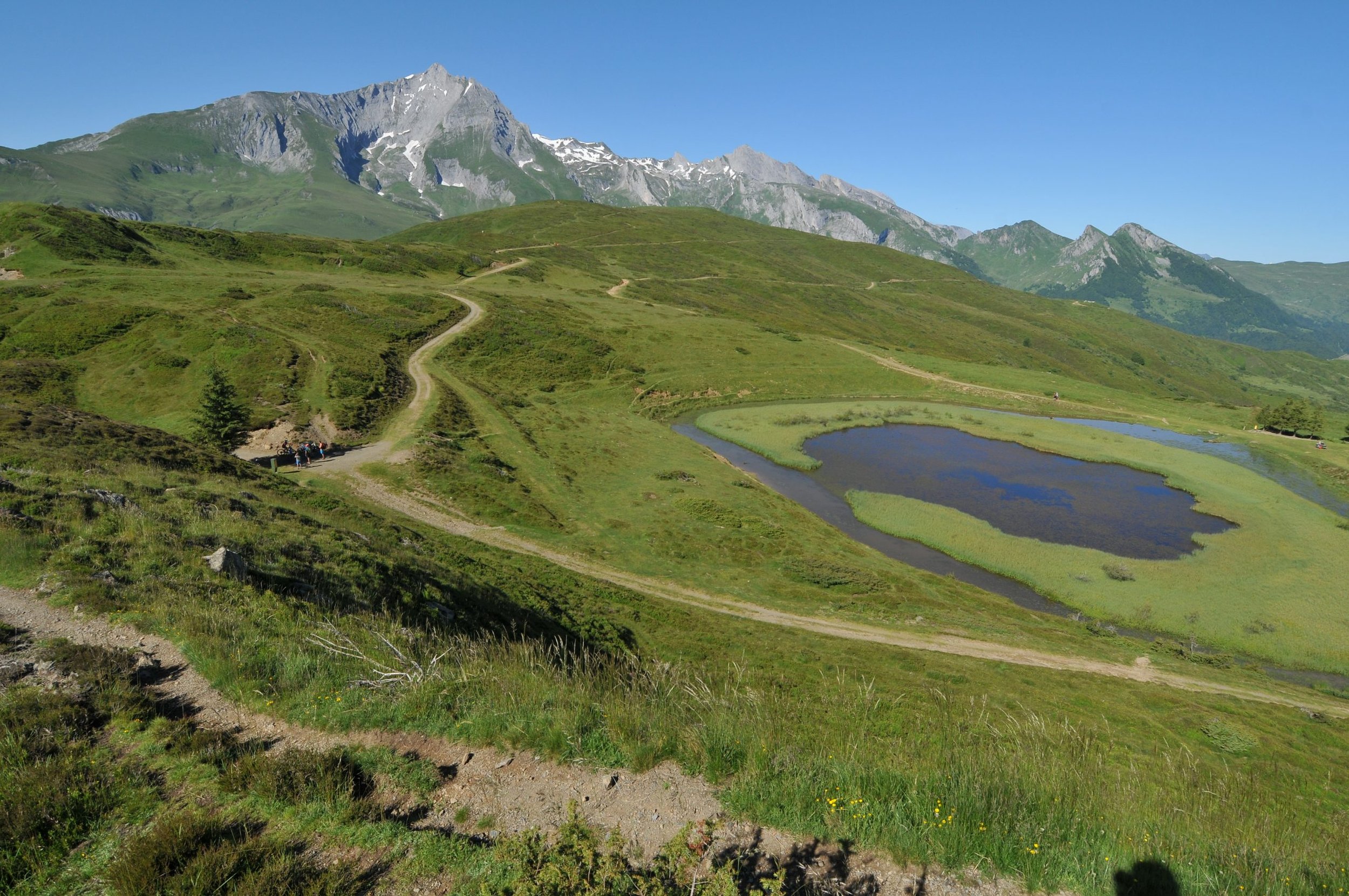 Paysage de montagnes avec des collines verdoyantes, un lac, un sentier serpentant et un ciel bleu clair.