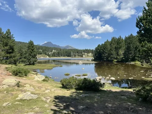 Paysage de lac entouré d'arbres avec montagnes en arrière-plan et ciel partiellement nuageux.