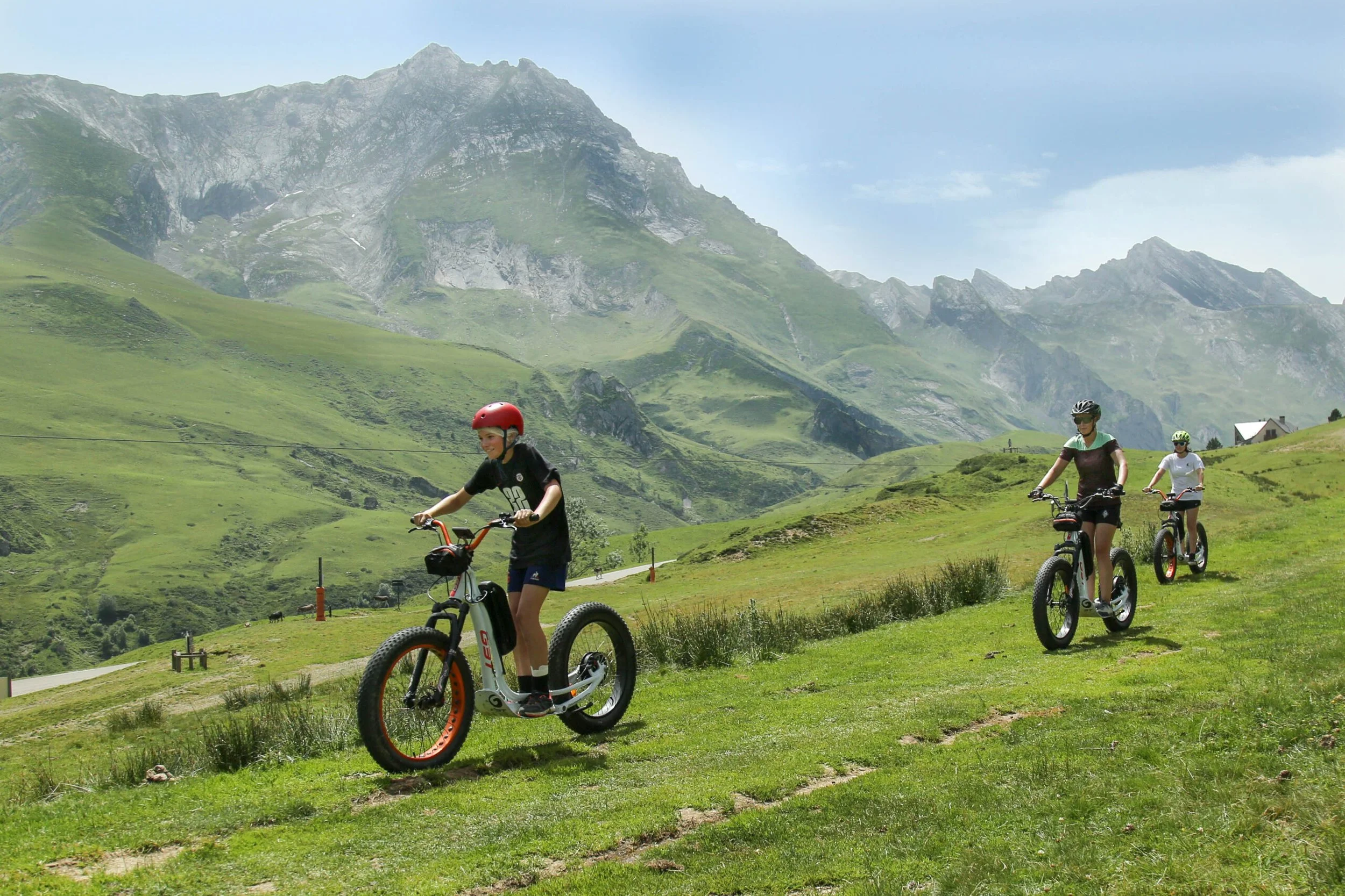 Trois personnes faisant du vélo électrique en montagne verdoyante avec des montagnes en arrière-plan sous un ciel en partie nuageux.