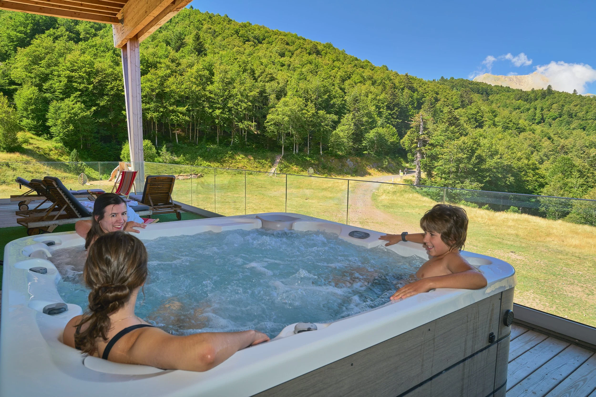 Groupe d'enfants et une femme profitant d'un jacuzzi sur une terrasse avec vue sur une forêt et des montagnes en arrière-plan, sous un ciel ensoleillé.