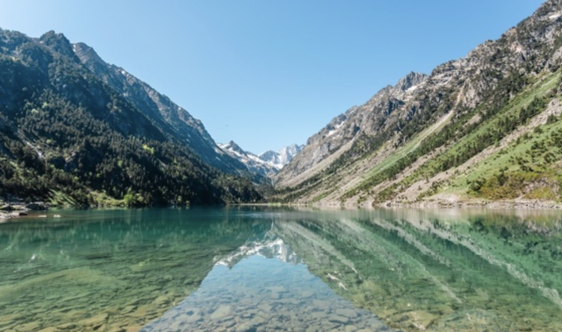 Lac vert entouré de montagnes rocheuses avec des sommets enneigés sous un ciel bleu clair.