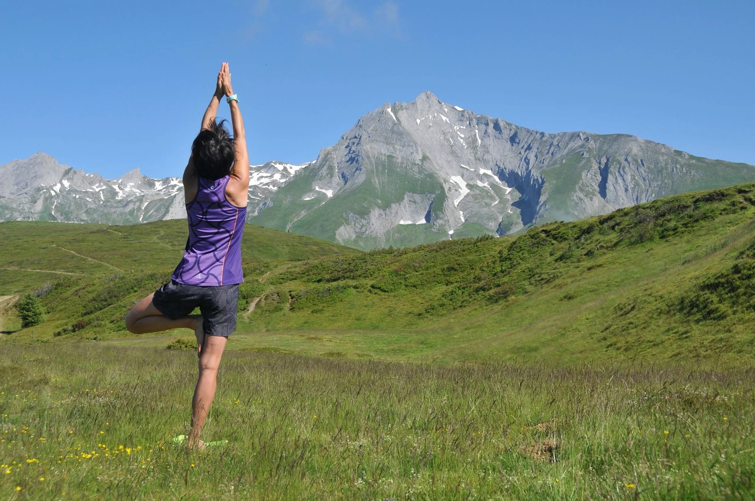 Une femme pratiquant le yoga dans un champ vert avec des montagnes en arrière-plan par beau temps.