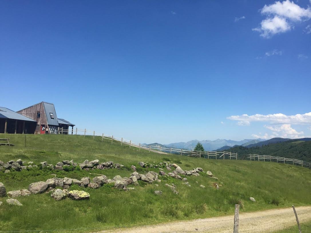 Paysage rural avec une maison moderne en bois, un mur de pierres, une clôture en bois, un sentier, et des montagnes en arrière-plan sous un ciel bleu avec quelques nuages.