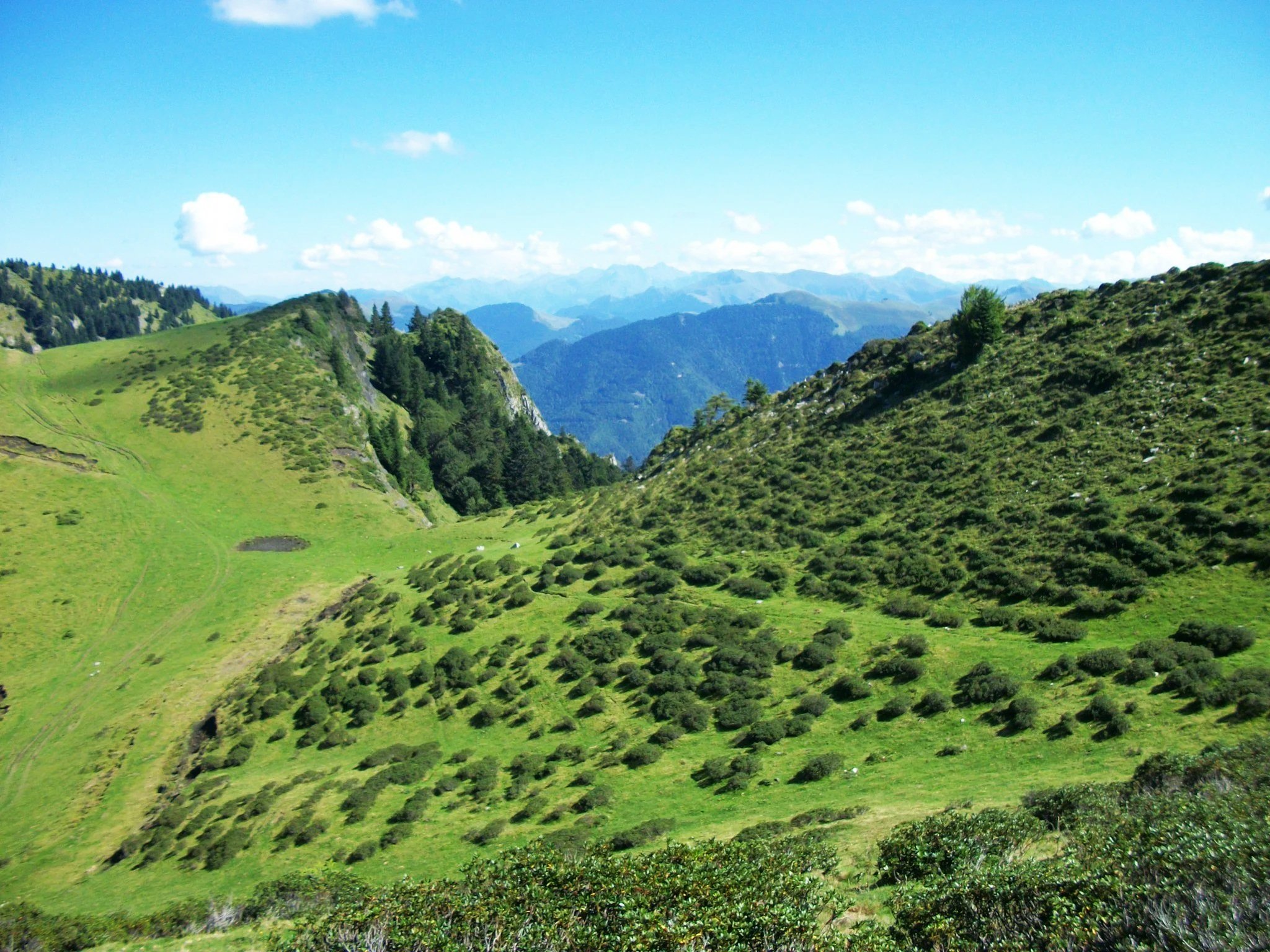 Paysage de montagnes verdoyantes avec collines escarpées, arbres et ciel bleu avec des nuages blancs.