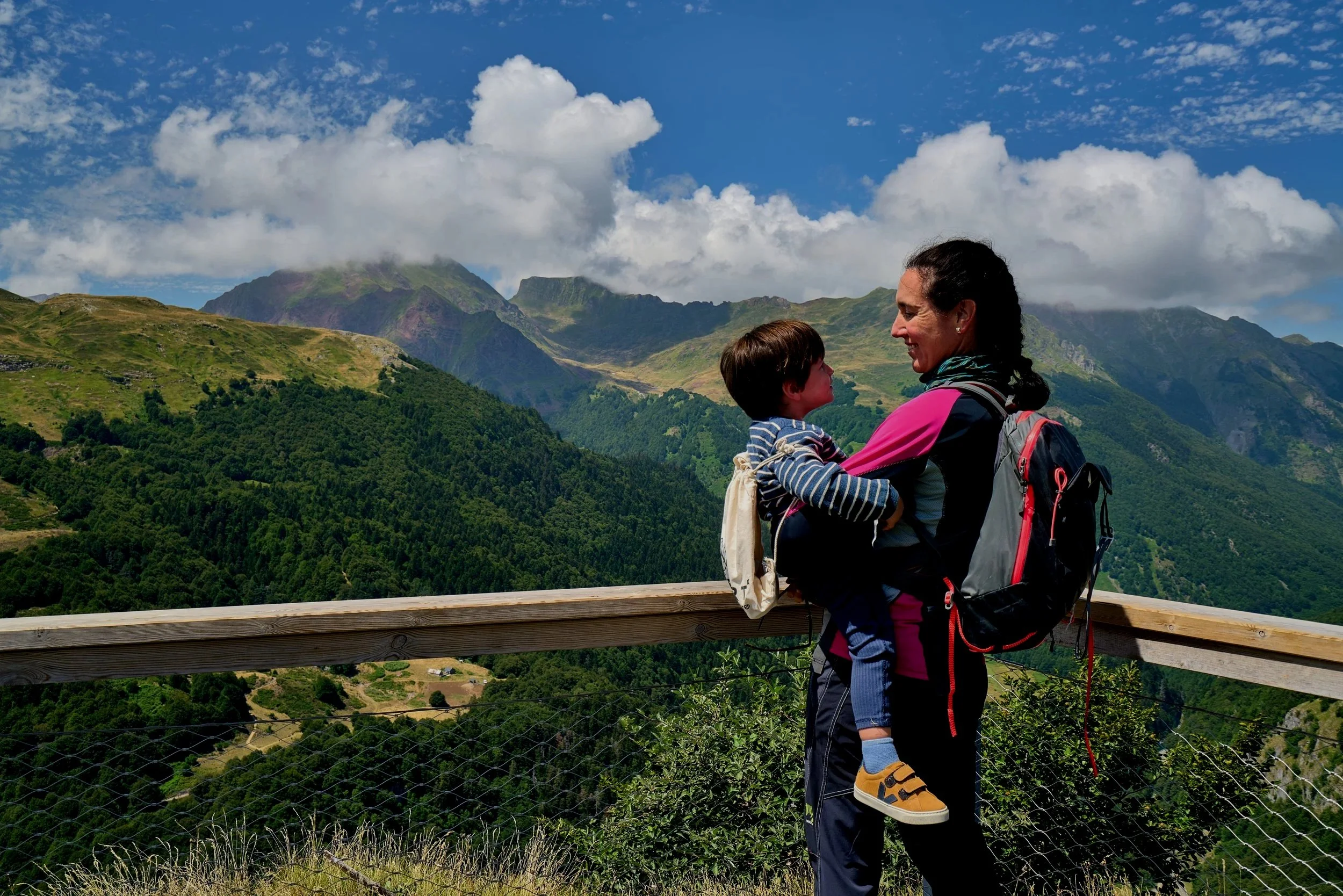 Une femme et un enfant regardent vers le paysage de montagnes verdoyantes sous un ciel partiellement nuageux, ils sont debout derrière une barrière en bois sur un sentier de randonnée.