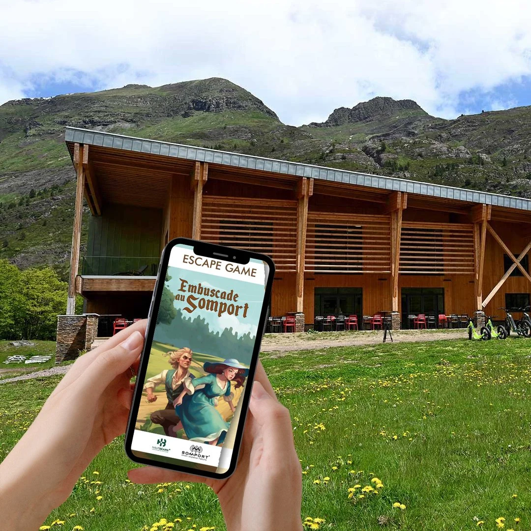 Une personne tient un smartphone affichant le jeu Escape Game Embuscade au Somport, avec une bâtisse en bois et des montagnes en arrière-plan.