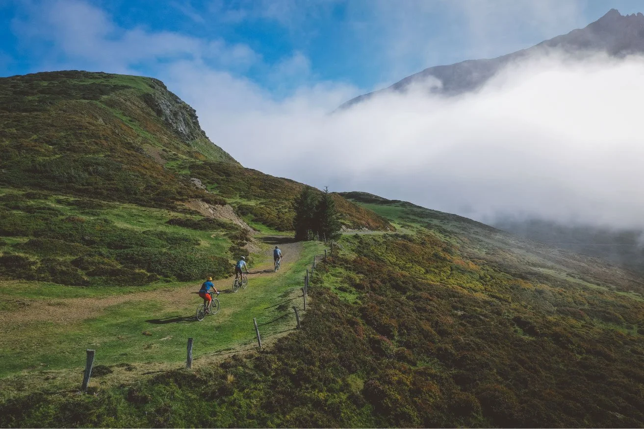 Trois personnes font du vélo sur un sentier de randonnée dans une vallée montagnarde, entourée de collines verdoyantes et de brouillard.