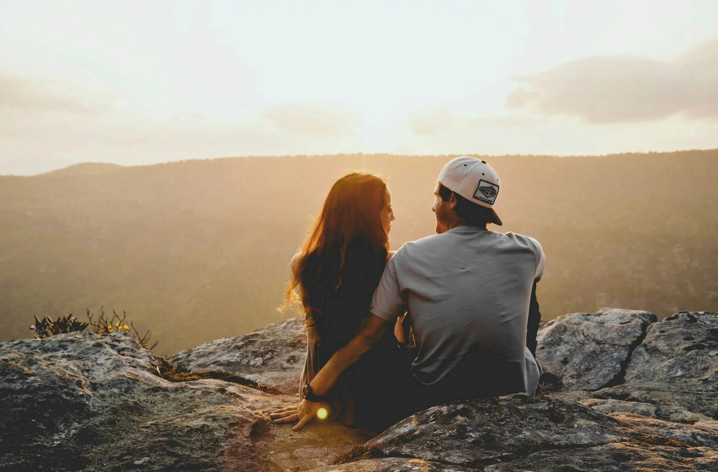 Un couple assis sur un rocher au sommet d'une montagne, regardant le coucher de soleil sur un paysage vallonné.