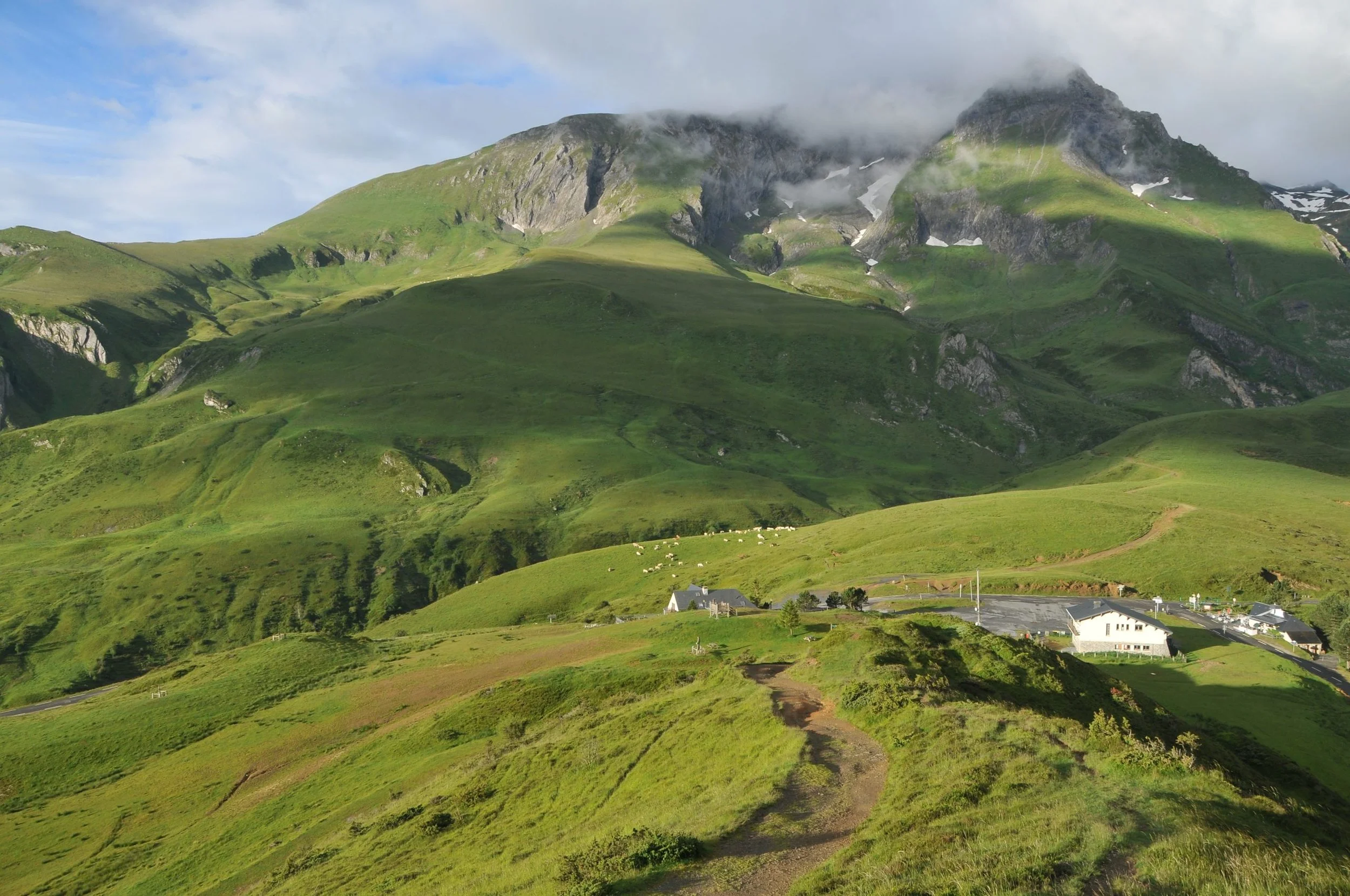 Paysage de montagnes vertes avec des nuages recouvrant le sommet, pâturages et quelques bâtiments en bas.