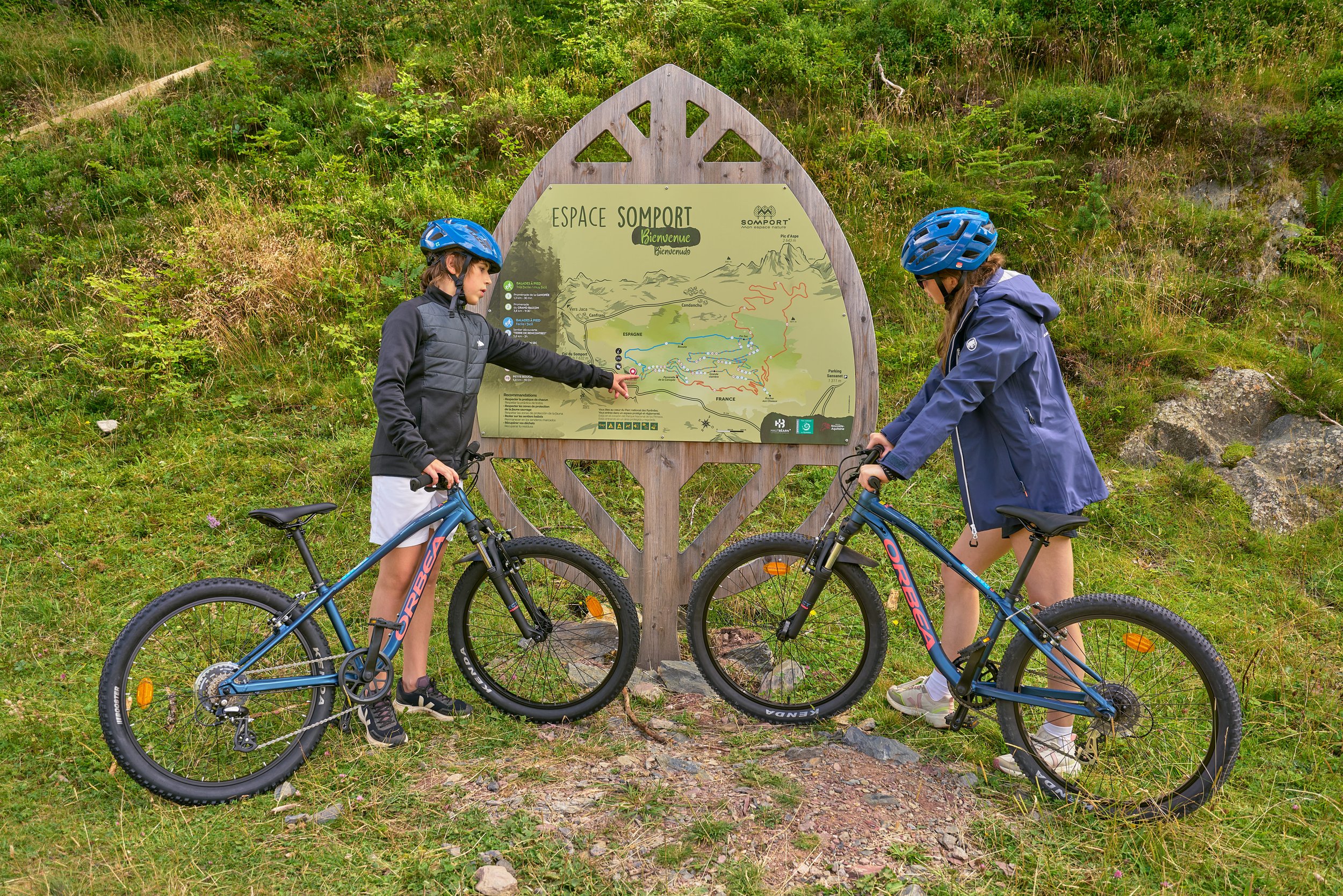 Deux jeunes filles avec des casques de vélo discutent devant un panneau d'information dans la nature, avec des vélos à côté d'elles.