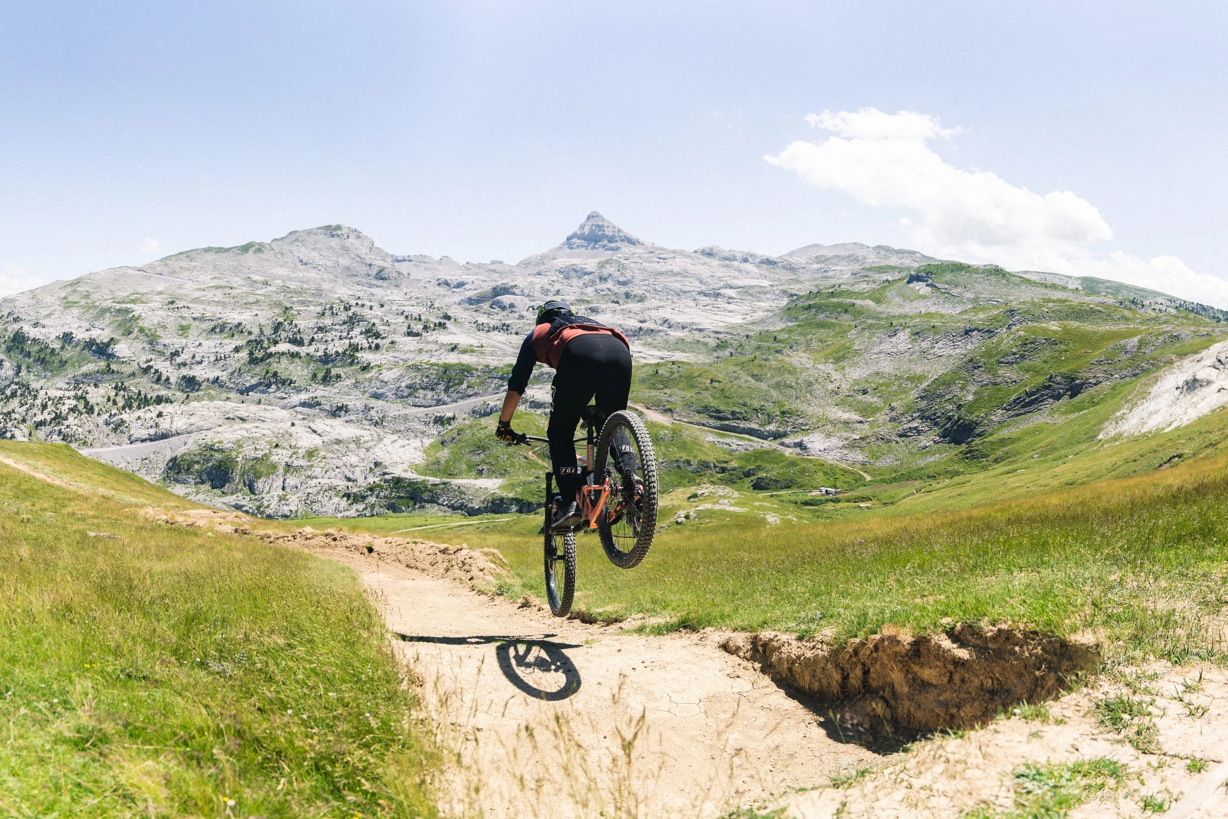 Vélo de montagne avec un cycliste effectuant un saut sur un sentier rocheux dans un paysage de montagnes vertes et rocheuses sous un ciel nuageux