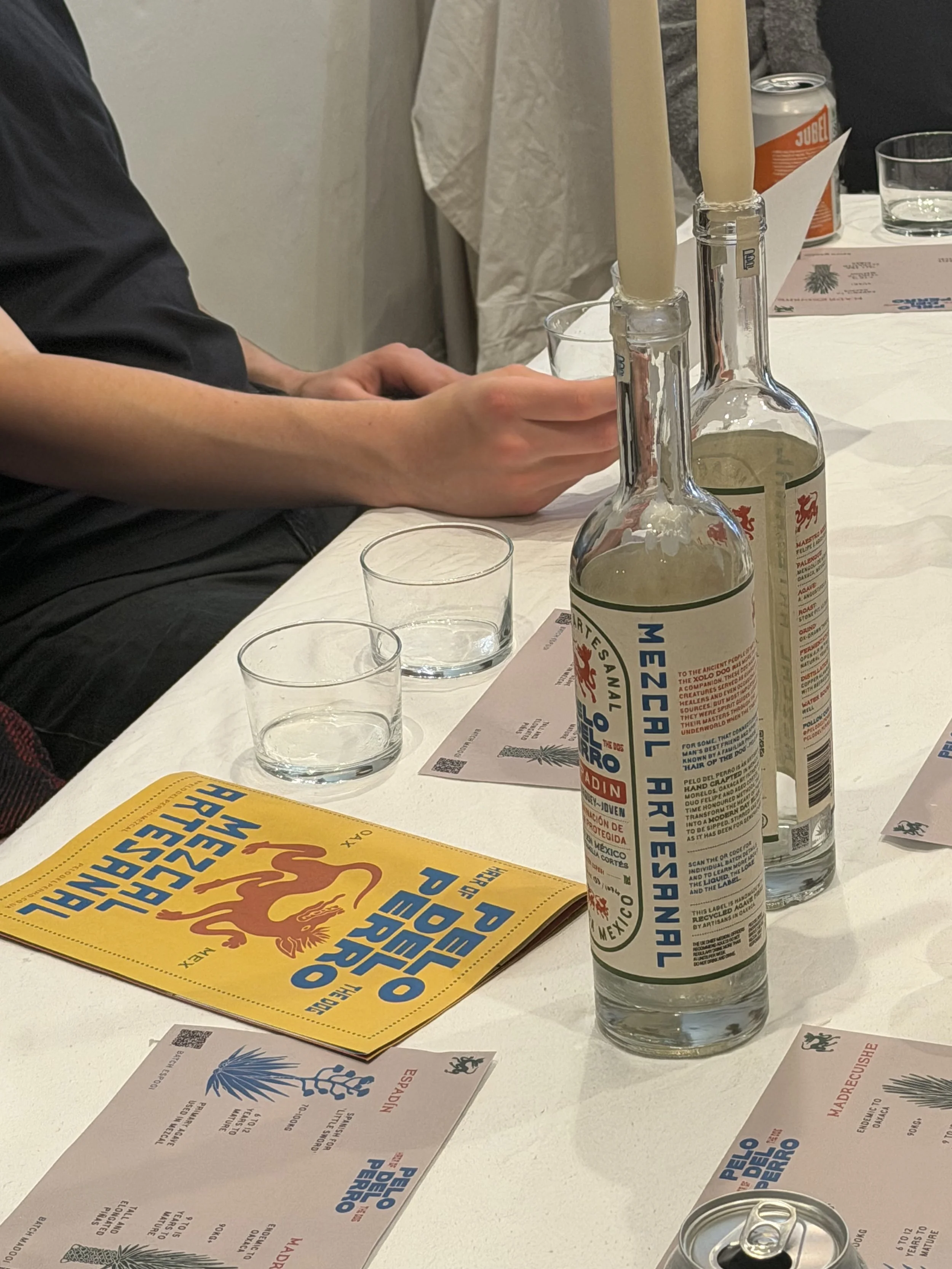 Table setting with two candles in glass holders, several empty glasses, a yellow booklet titled 'PERRO AZUL MÉXICO,' and informational cards in Spanish, with a partially visible soda can and a glass of water.