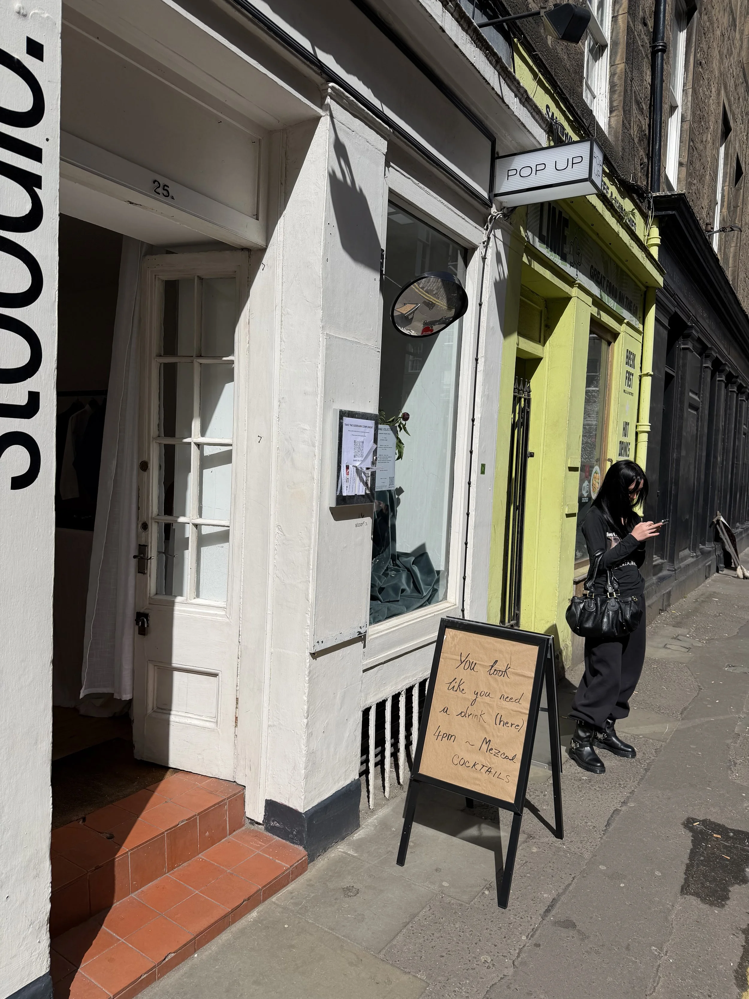 A person standing outside a storefront, looking at their phone. The storefront has a sign that reads 'POP UP' and a chalkboard sign with a handwritten message offering drinks and cocktails. The building is painted in light green and black, with a large glass window revealing the interior.