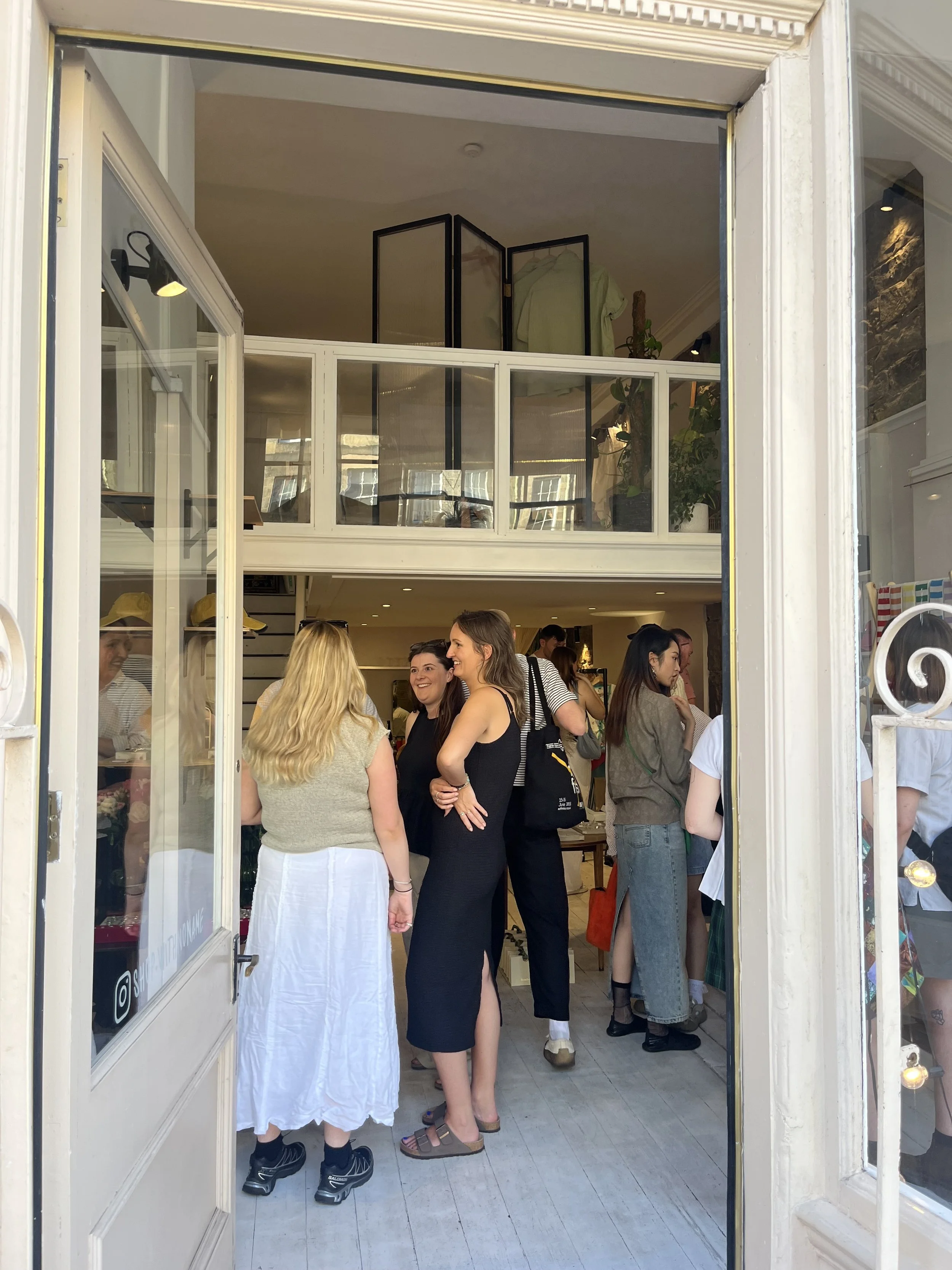 People gathered outside a storefront with a sign that says 'Ludic Studio'. The shop has a large window displaying a plant and writing utensils, with a neon sign that reads 'Reservation'. The building has a brick facade with two upper windows and some hanging wires, and the street has cobblestone paving.