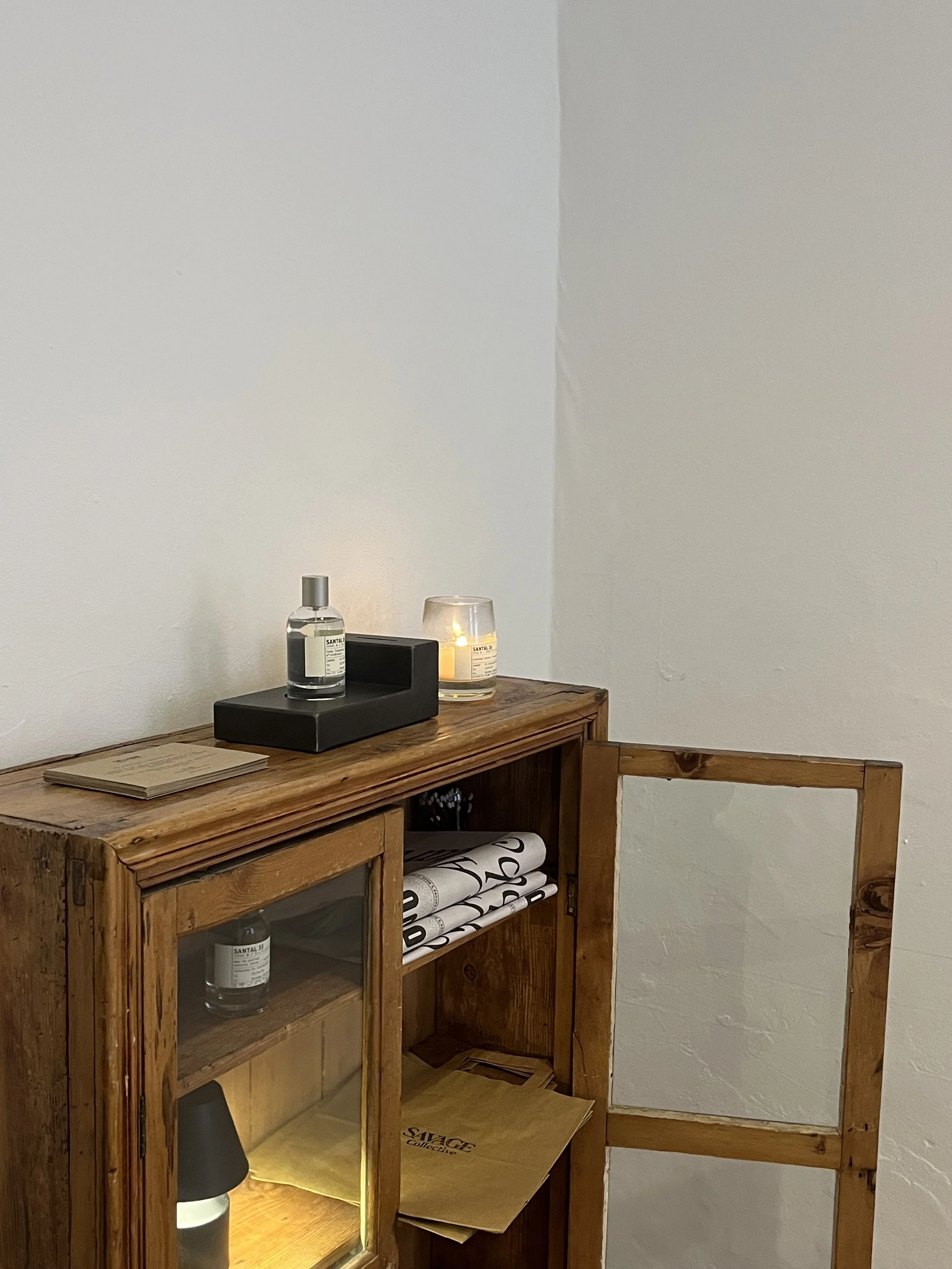 A small wooden cabinet with glass doors, a spray bottle, a candle, and some rolled paper inside, positioned against a plain white wall.
