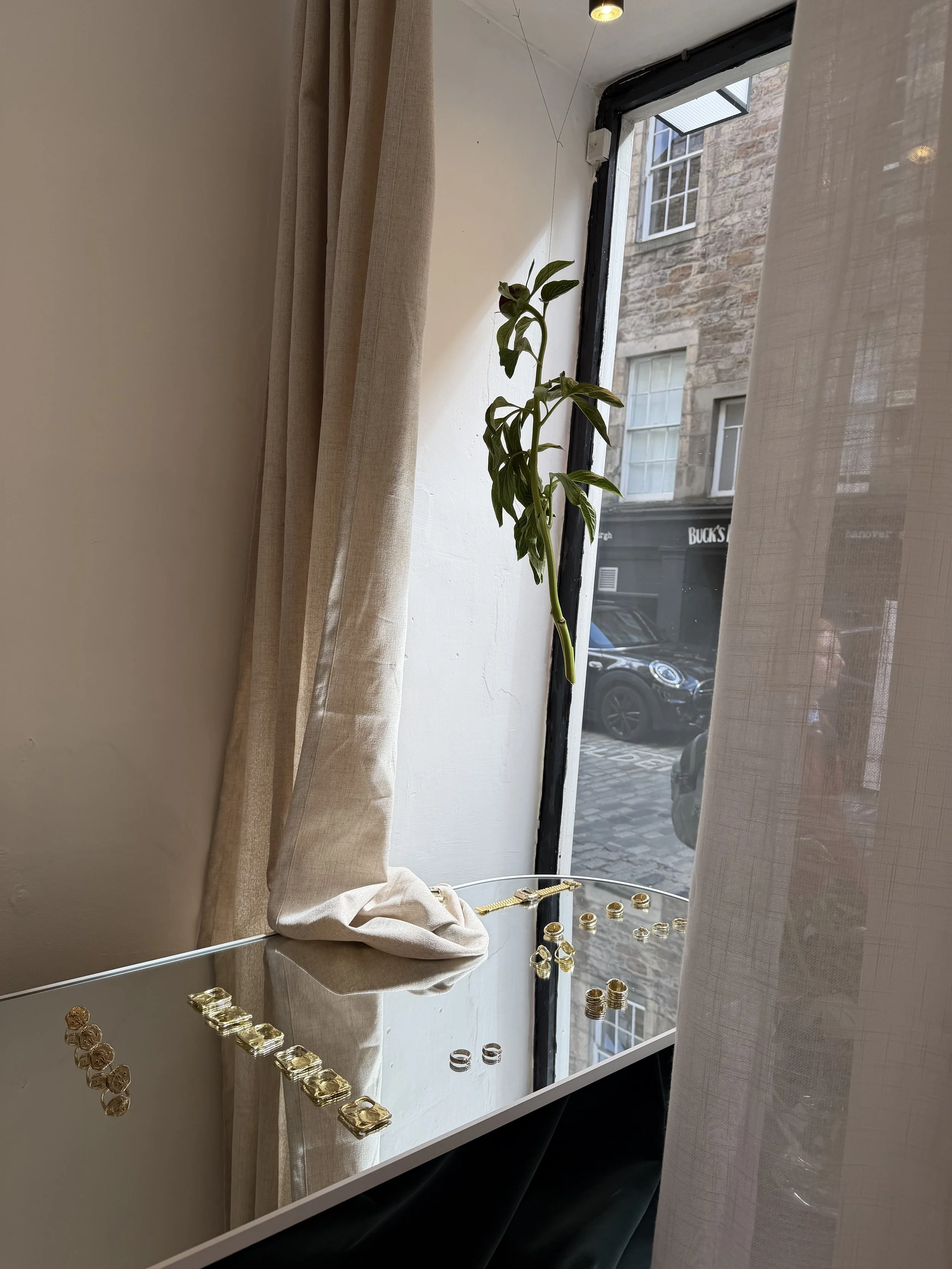 A display of shiny gold and silver rings on a white surface outside a shop window with beige curtains and a view of a cobblestone street and brick building outside.