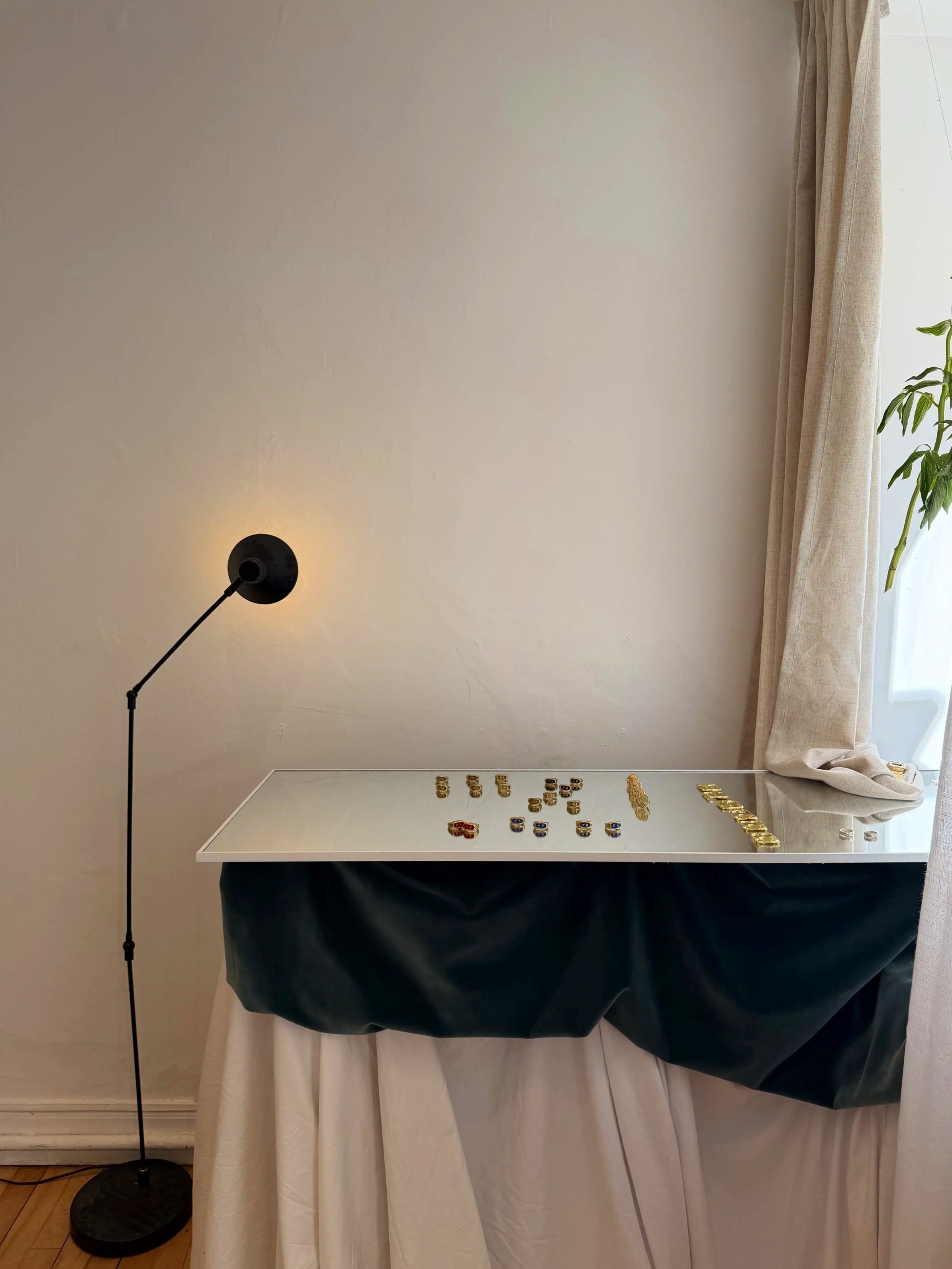 A white table with a collection of poker chips arranged on it, a black tablecloth underneath, a minimalist black floor lamp to the left, white curtains to the right, and a partially visible window with natural light.