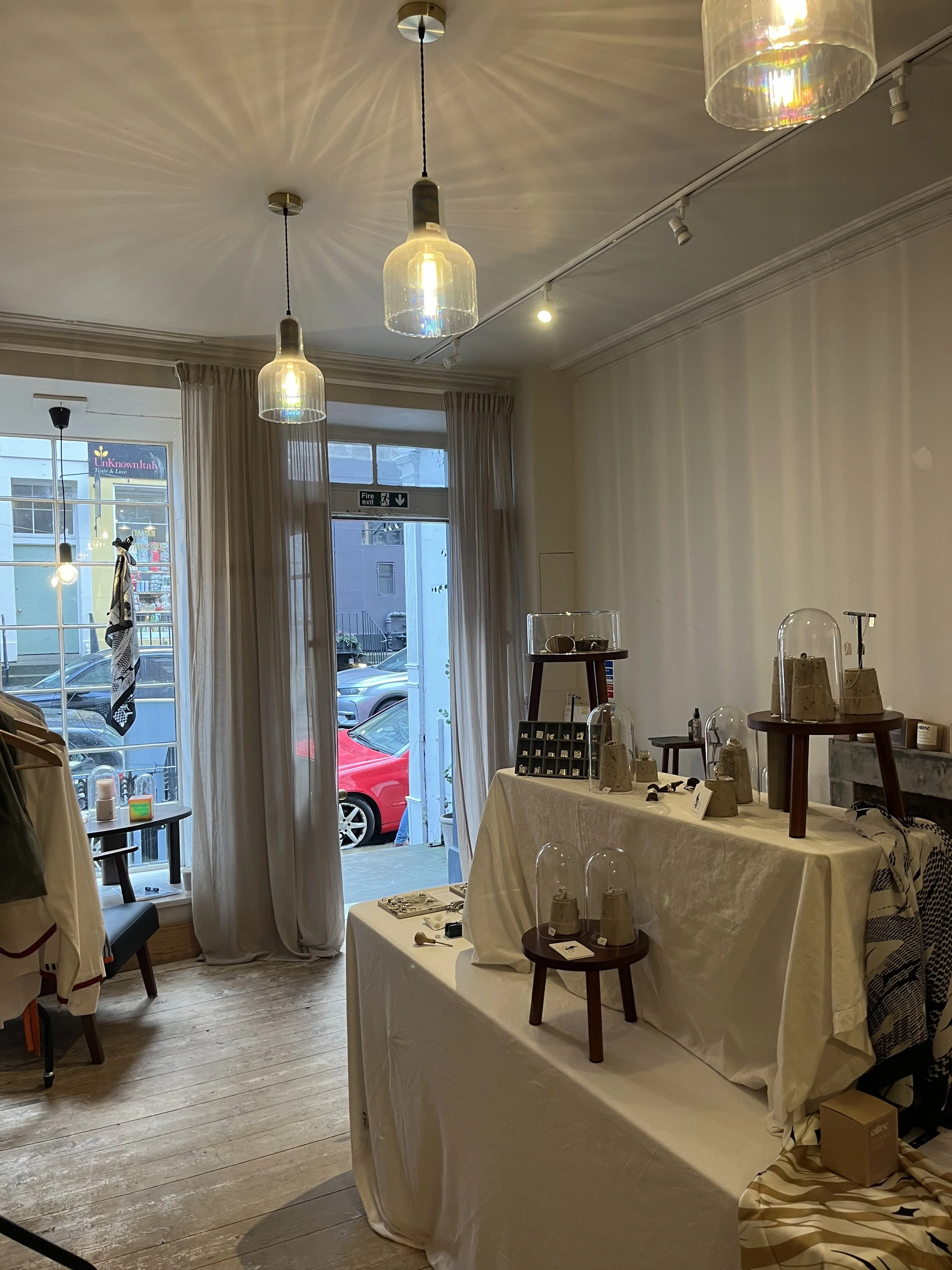 Interior of a store with hanging light fixtures, tables displaying various items, and a large window with curtains. Cars can be seen outside through the window.