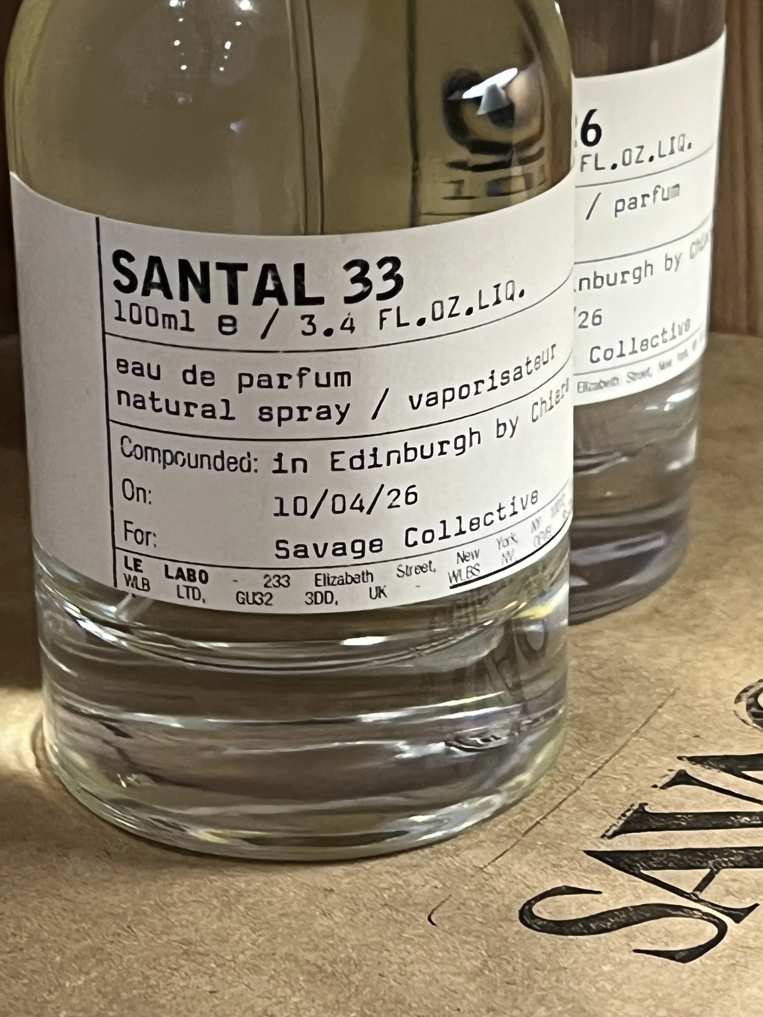 Close-up of a perfume bottle with a white label, displaying the brand Santal 33 in bold black letters, placed on a wooden surface with black handwritten text partially visible.