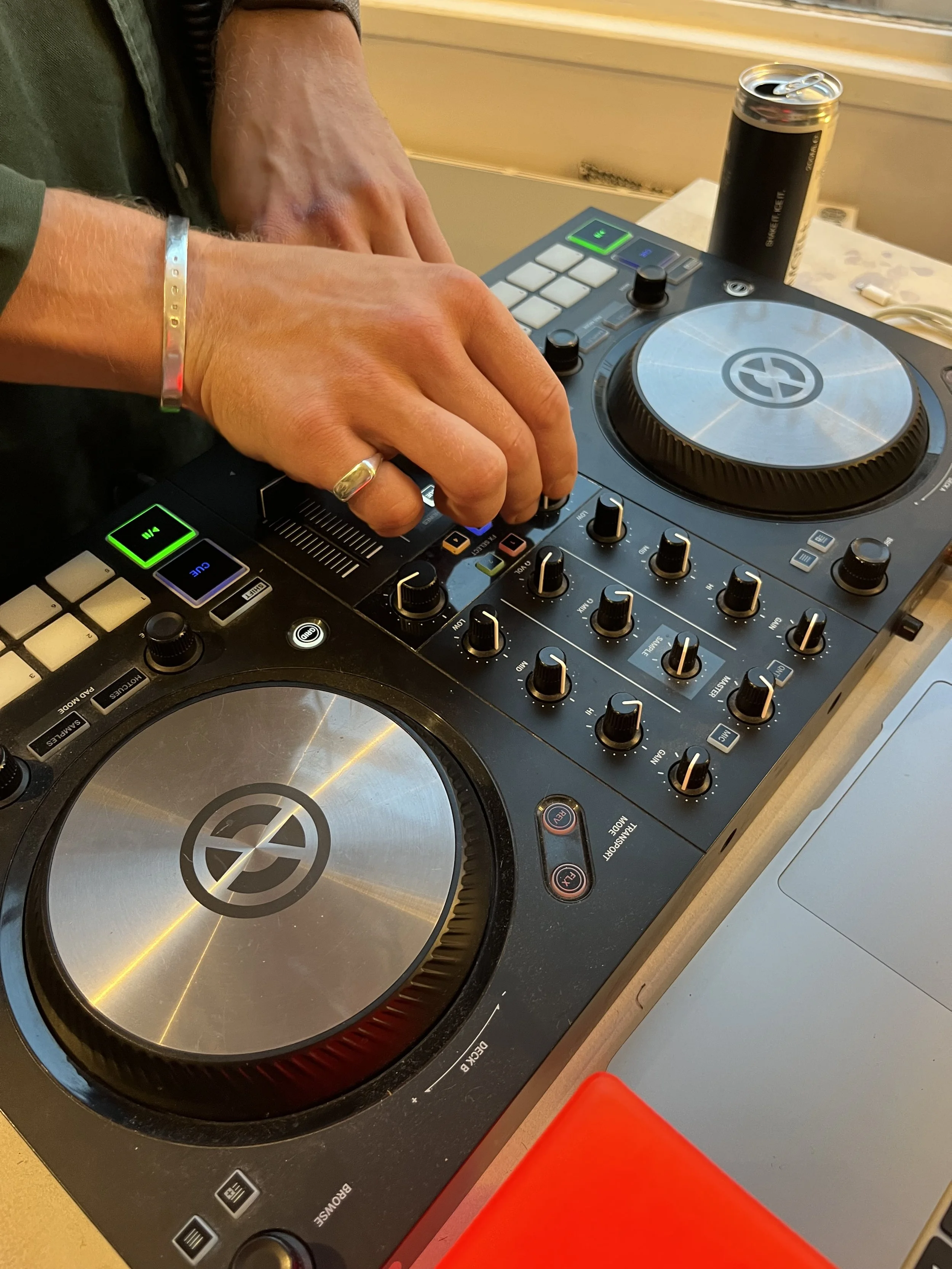 Close-up of a person's hands operating a DJ controller with two large jog wheels, various knobs, and buttons, a can of Red Bull energy drink in the background, and a red object at the bottom edge.