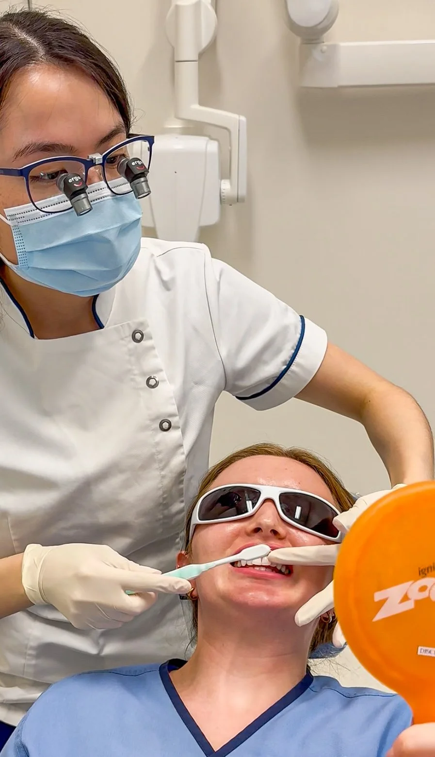 A dentist wearing magnifying glasses and a face mask is examining a patient’s teeth who is wearing protective sunglasses during a dental check-up.
