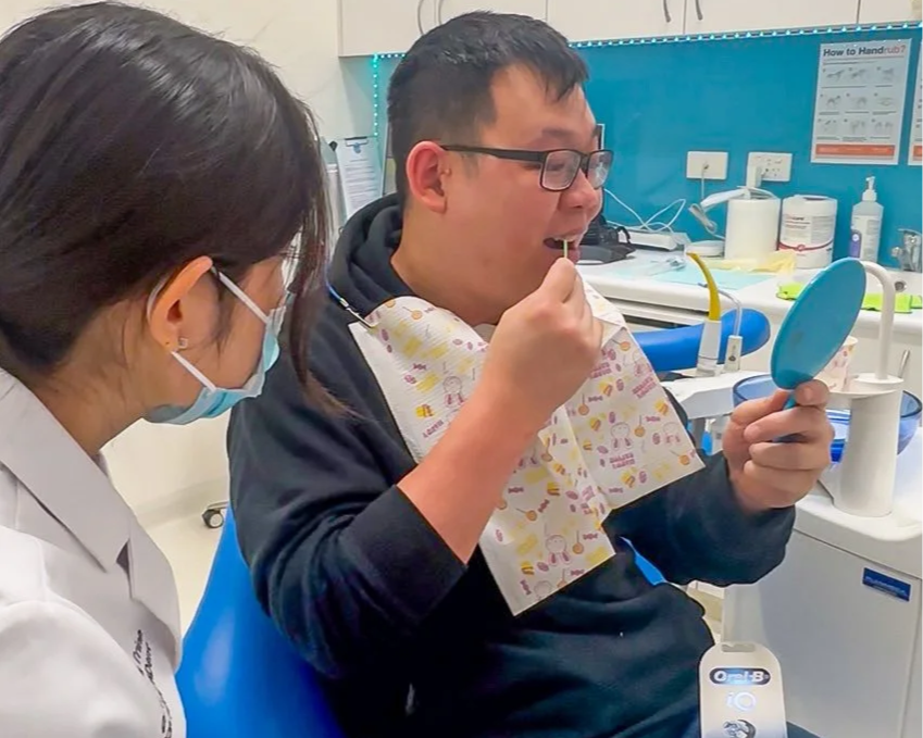 A man in glasses and a dental bib looking into a small mirror and applying lipstick while sitting in a dental office, with a dental professional nearby.