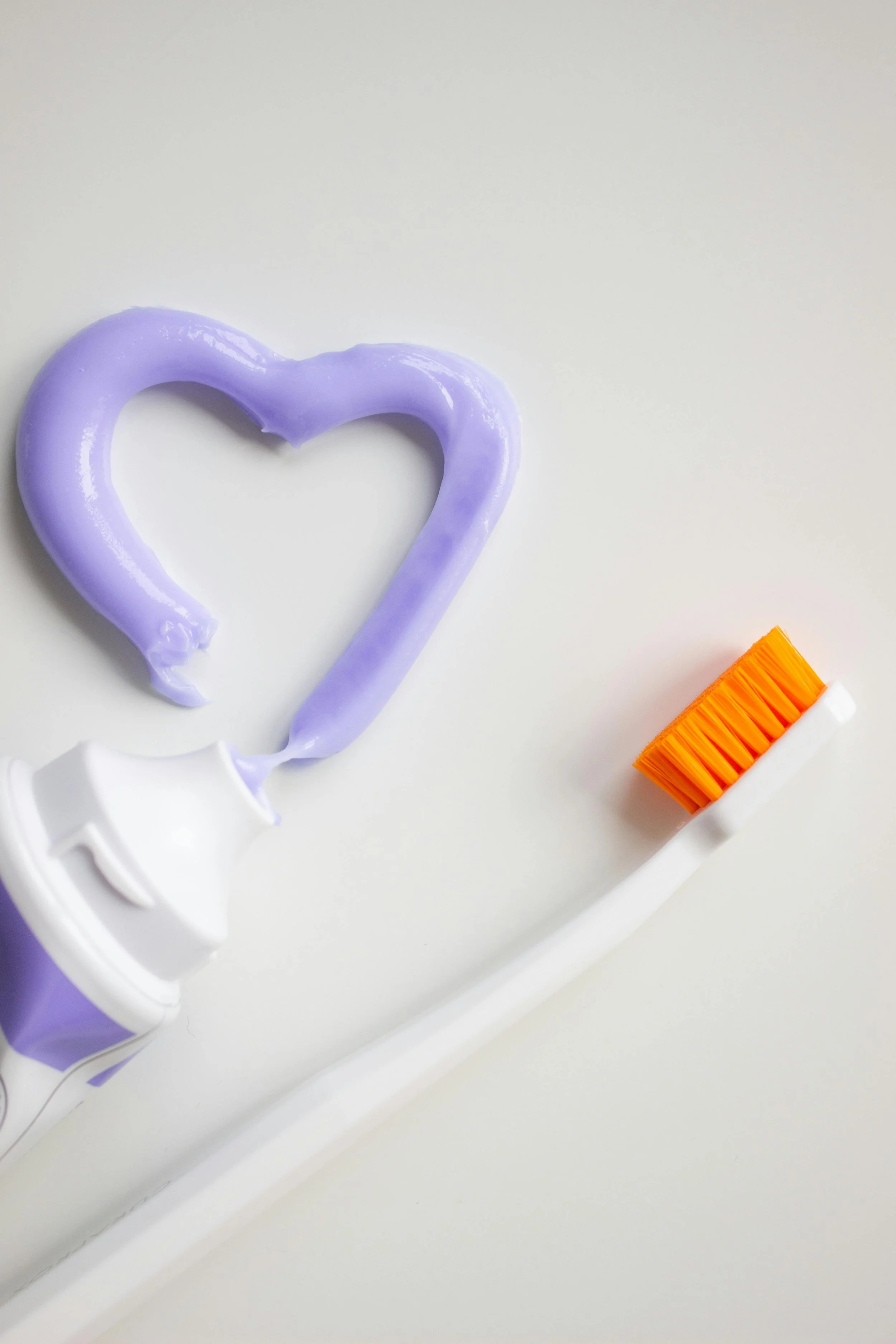 A heart shape is made with purple lipstick on a white background. A white tube of lipstick with an orange brush is partially visible at the bottom.