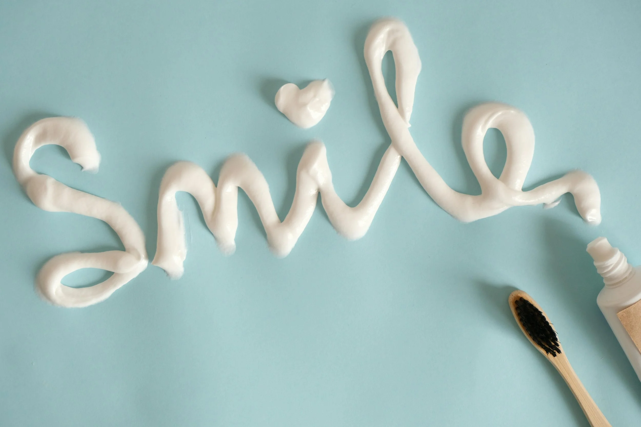 Creamy white substance spelled out on a light blue background, with a toothbrush and a small container of black mascara or eyeliner nearby.