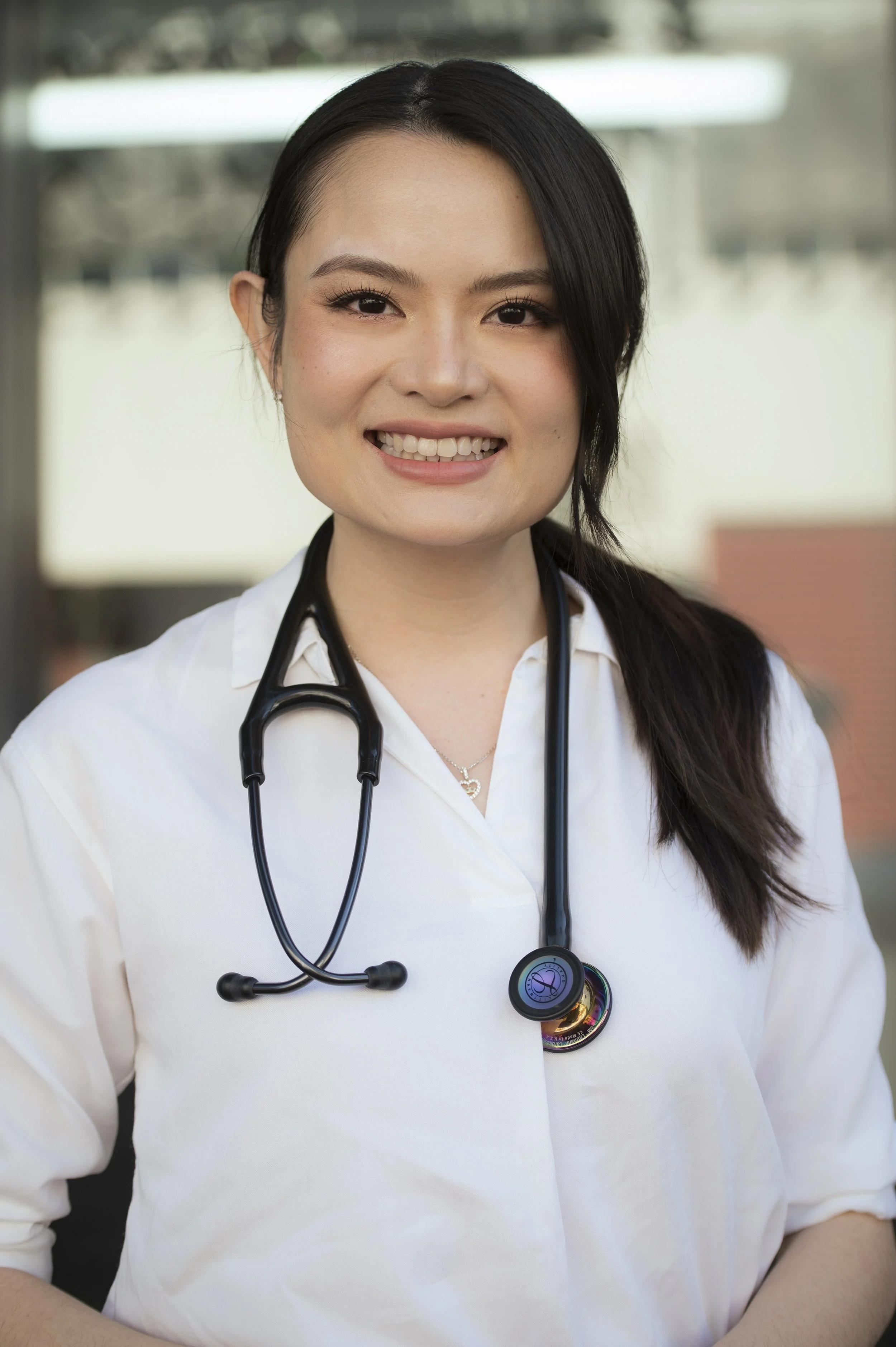 Close-up of a smiling female healthcare professional wearing a white uniform with a stethoscope around her neck. The background is blurred, suggesting an outdoor or clinical setting.