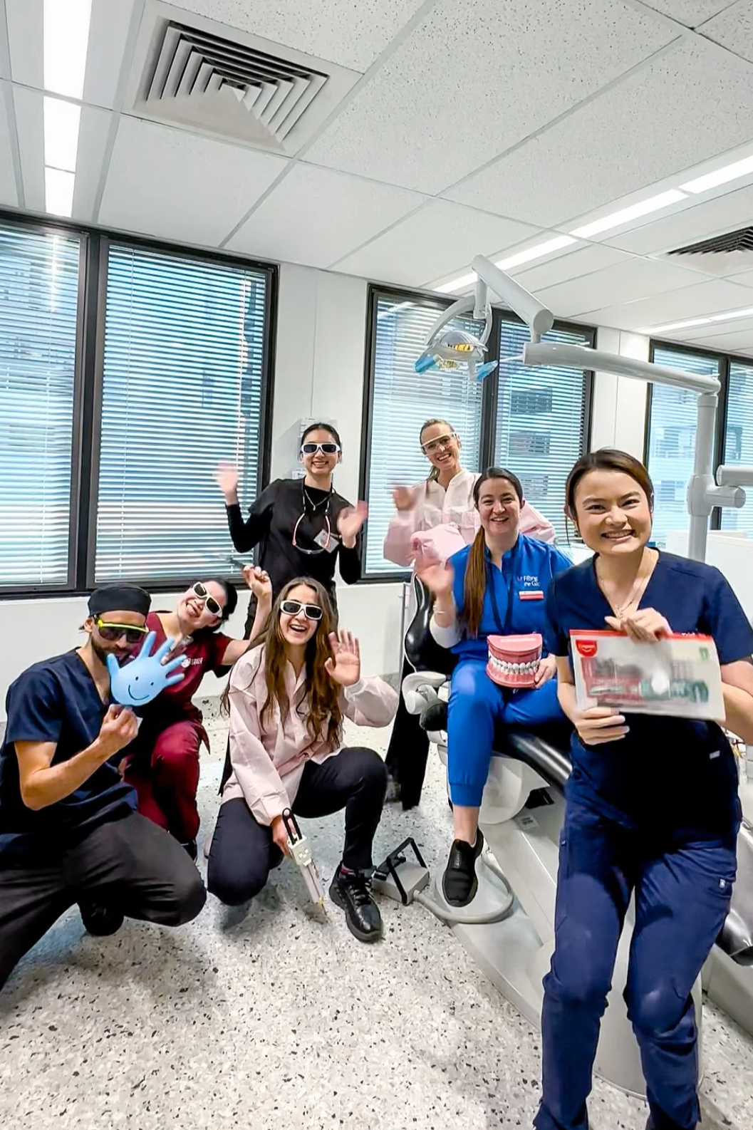 Group of dental professionals celebrating in a dental clinic, some wearing sunglasses and scrubs, holding dental tools and model teeth, smiling, and waving.