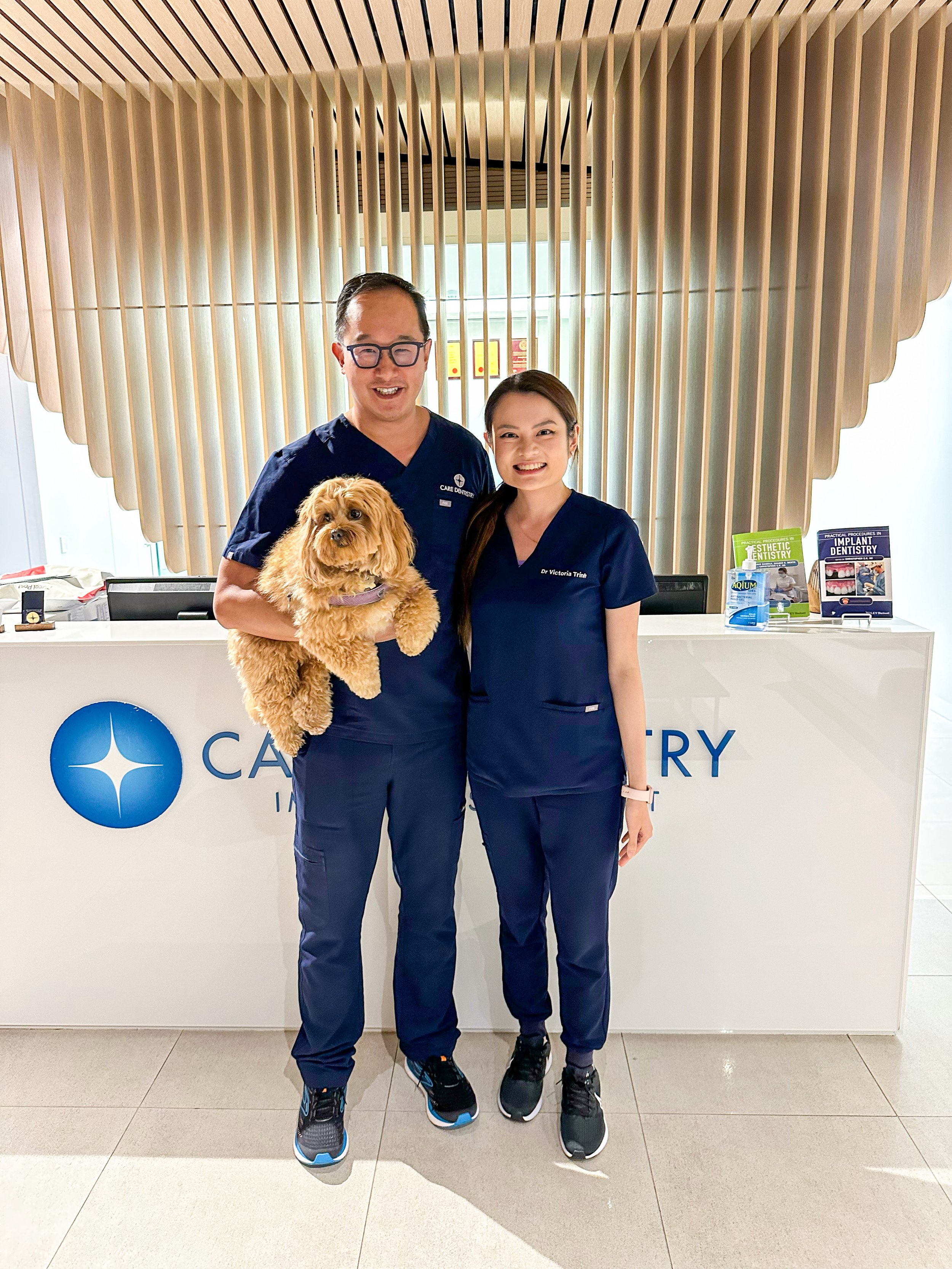 Two people, a man and a woman, standing at a reception desk, smiling at the camera, with a small fluffy dog being held by the man. The man is wearing glasses and navy scrubs, and the woman is wearing navy scrubs labeled 'Dr Victoria Trihi.' The reception desk has dental care informational pamphlets and a bottle of hand sanitizer. The background features wooden slats on the wall.
