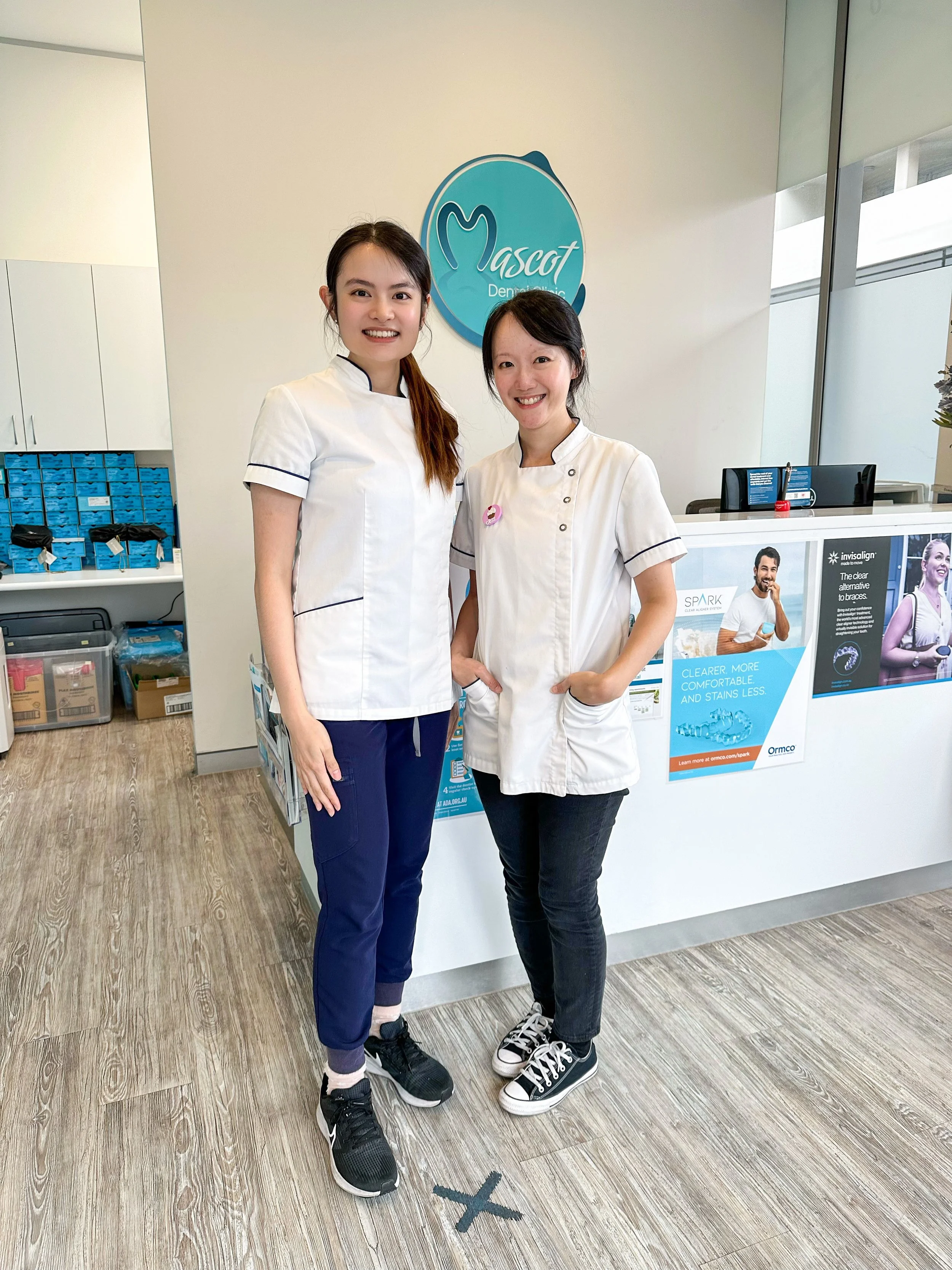 Two women standing inside a dental office, smiling at the camera. One woman is wearing a white medical uniform with dark blue accents, and the other is wearing a similar uniform with a pink badge. There is a reception desk behind them with promotional posters and a logo that says "Mascot Dental." The floor has a black X mark on it, indicating social distancing.