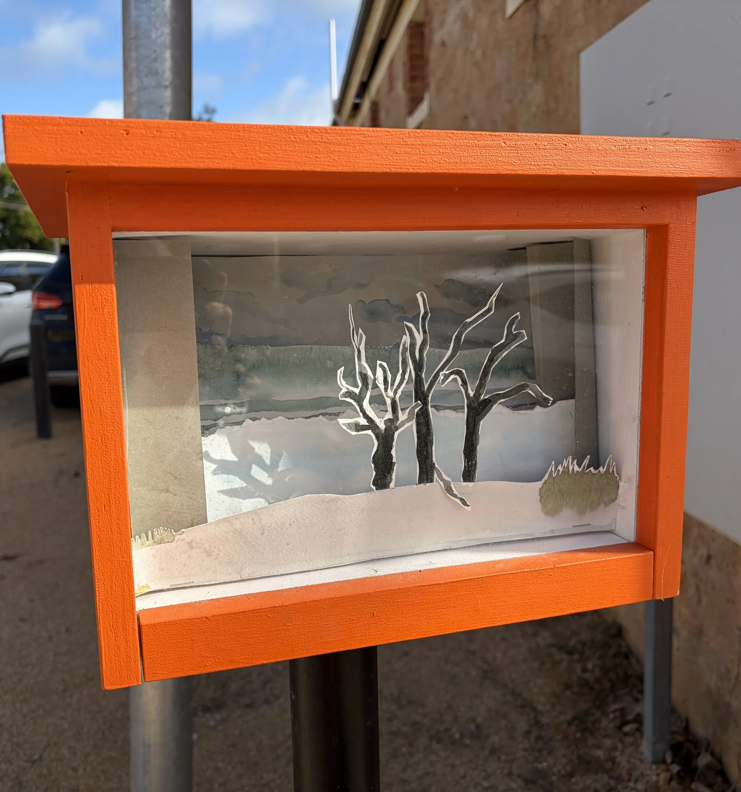 Wei Kari Calm watercolour installation depicting three bare trees beside still water beneath a soft, cloudy sky, displayed in a bright orange outdoor Pocket Gallery box in Waikerie.