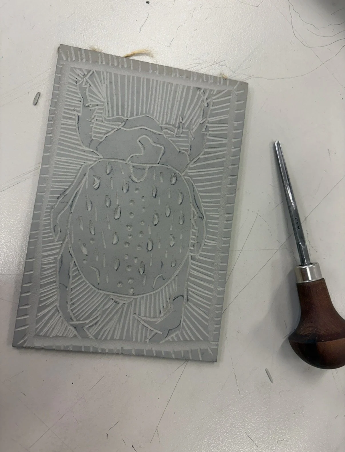 Hand-carved scarab beetle printing block with detailed linear patterns and carving tool beside it.