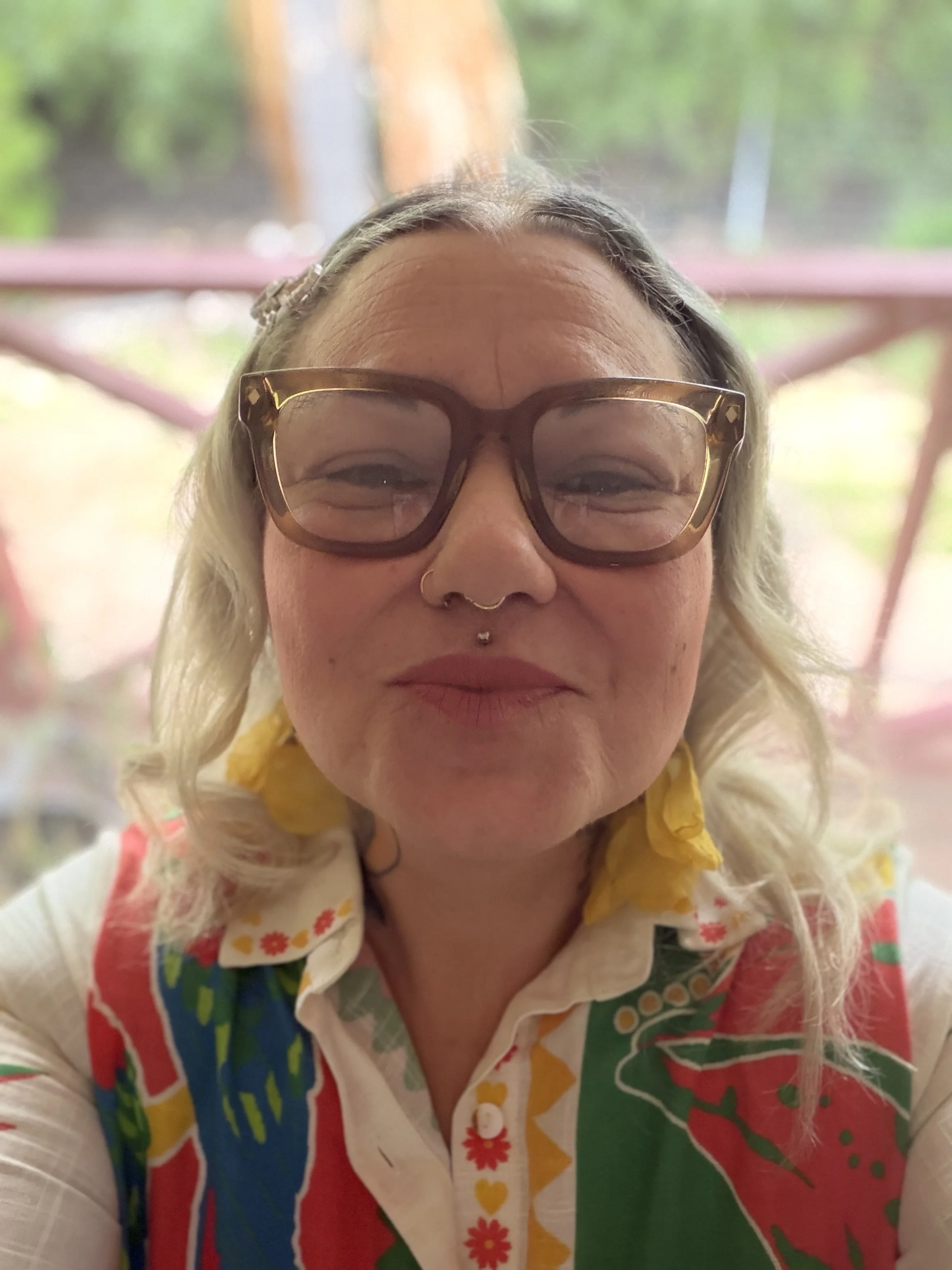Close-up of a woman with blonde hair wearing large tortoiseshell glasses and a colorful, patterned shirt, smiling and looking at the camera, outdoors with a blurry background.