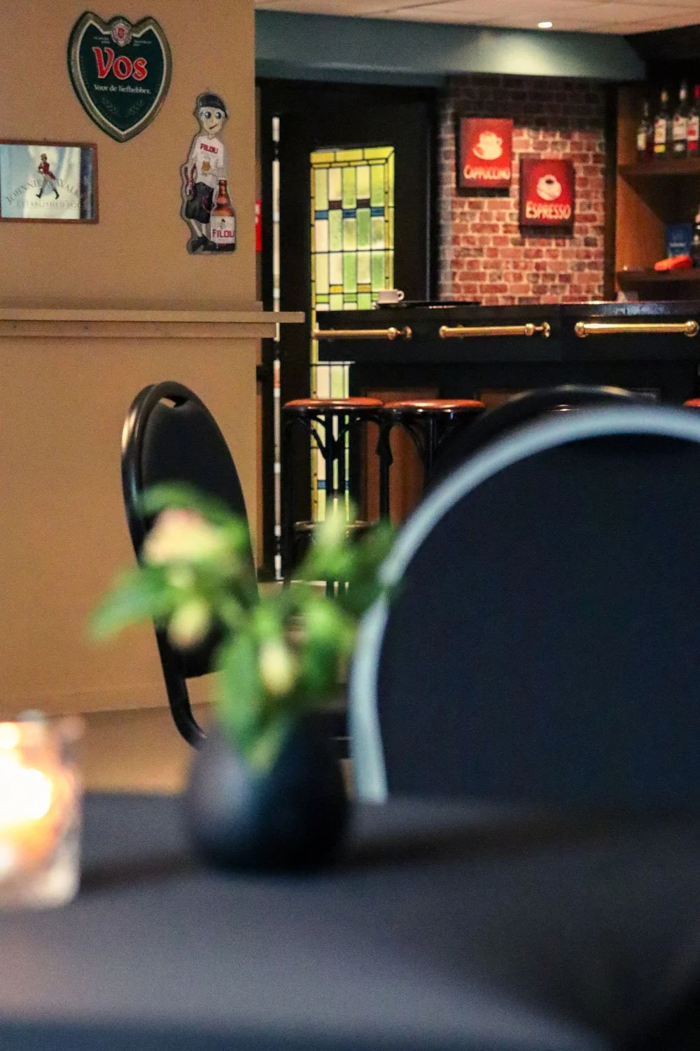 Interieur van een café of bar met een vage plant op een tafel, een muur met foto's en tekeningen, en een bar met krukken op de achtergrond.